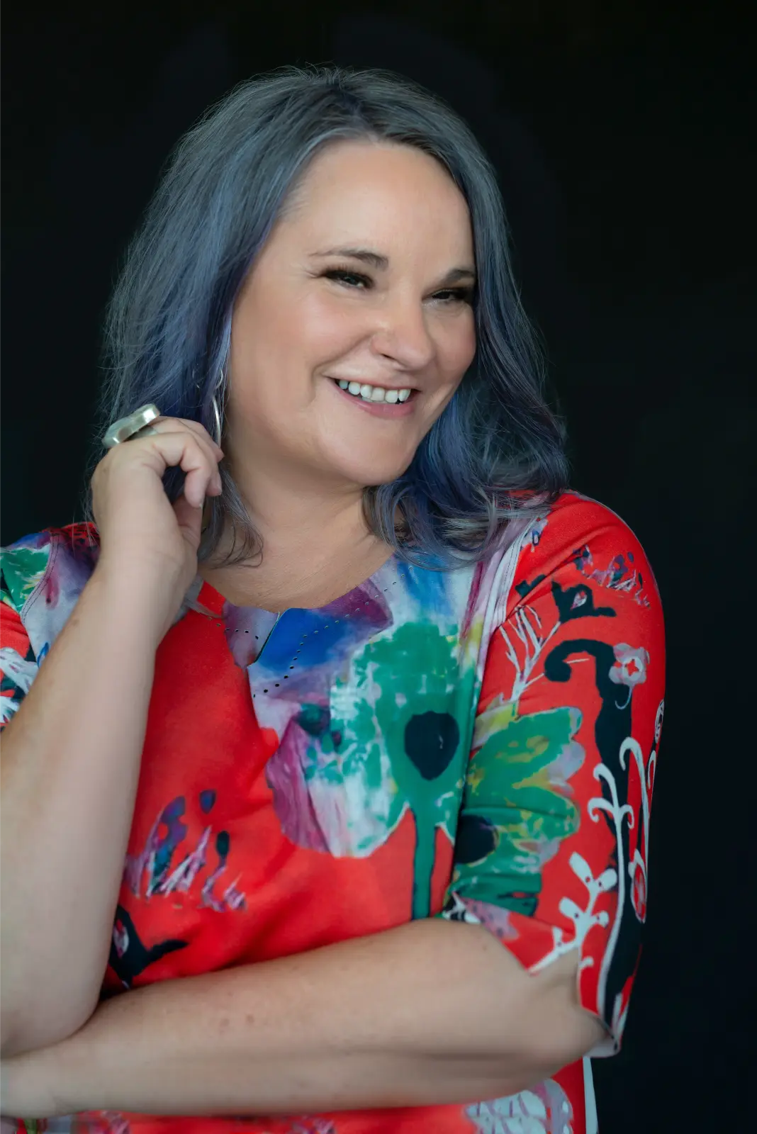 Smiling woman with shoulder-length blue-gray hair wearing a colorful, abstract-patterned red top against a black background.
