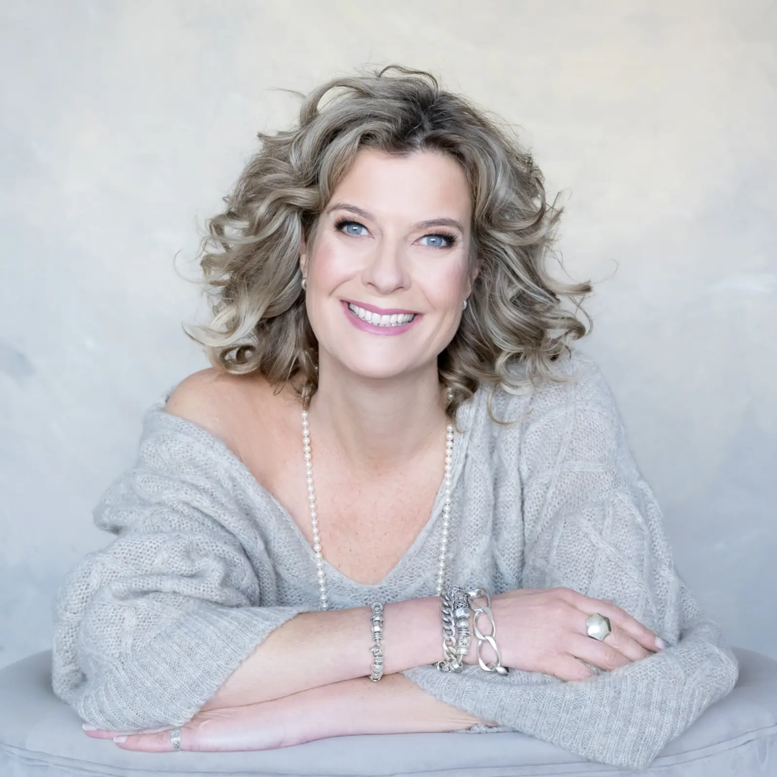 Smiling woman with curly blonde hair wearing a light gray off-shoulder sweater, pearl necklace, and silver bracelets, posed with her arms crossed on a cushioned surface against a light background.