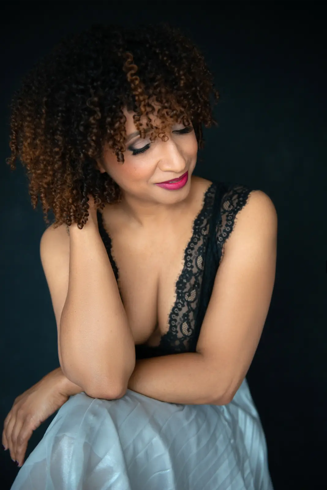 Woman with curly hair wearing a black lace-trimmed top and a light-colored skirt, looking down with a soft smile against a dark background.