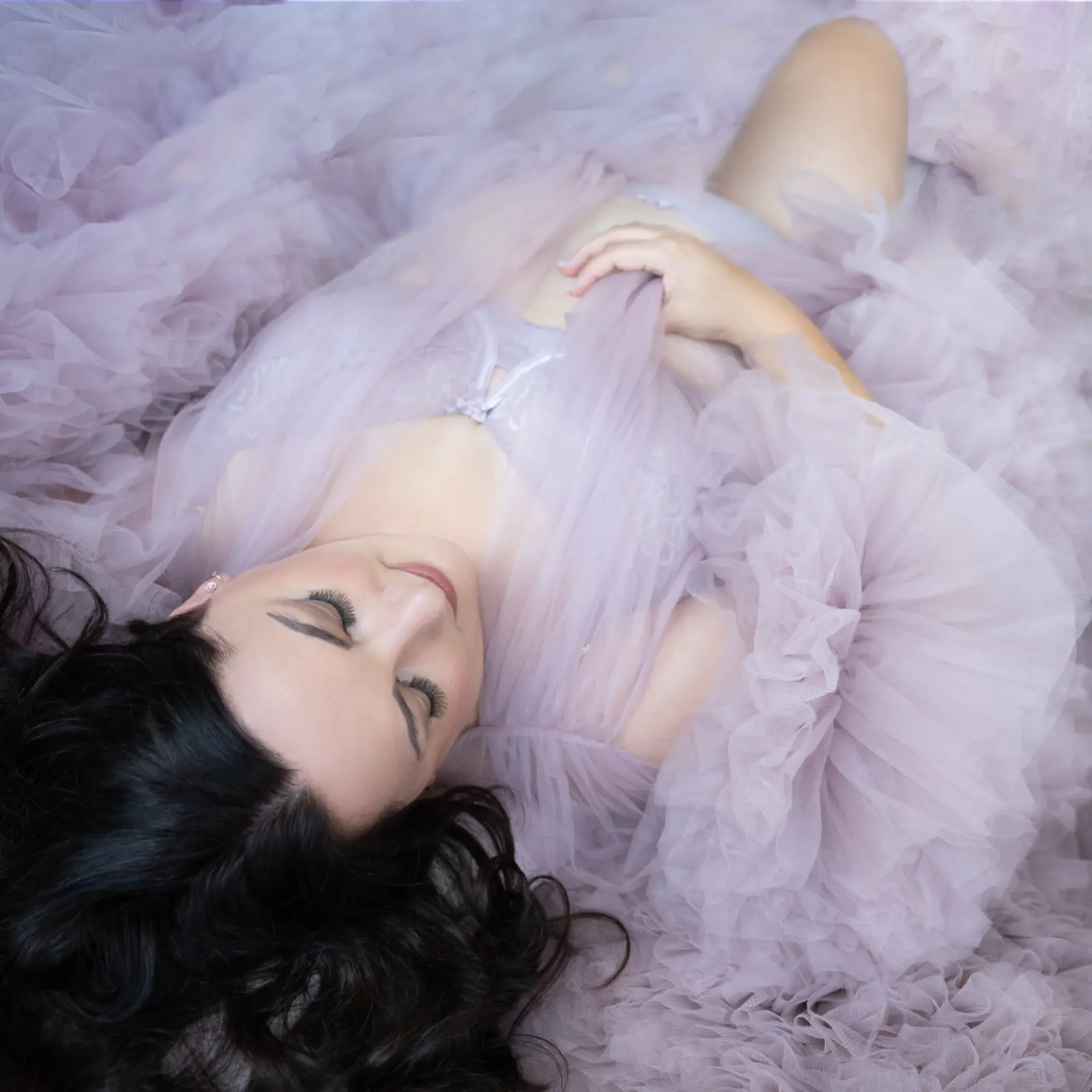 Woman with long dark hair lying down with eyes closed, wrapped in soft, flowing lavender tulle fabric.