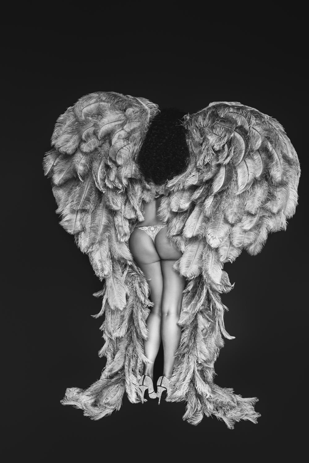 Black and white photo of a woman standing with her back to the camera wearing large feathered angel wings, a thong, and high heels, against a dark background.