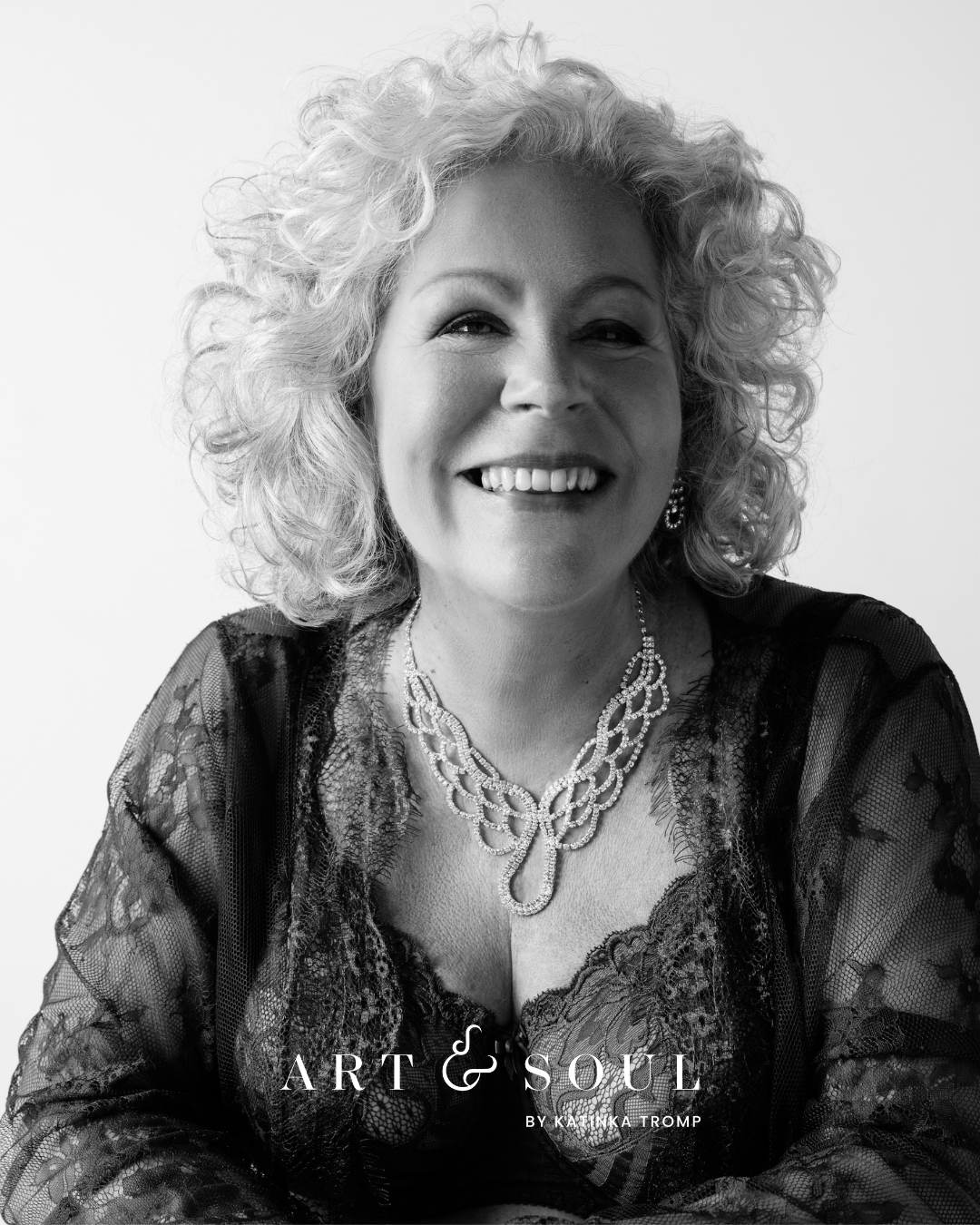 Black and white portrait of a smiling woman with curly hair wearing a lace top and an ornate necklace.