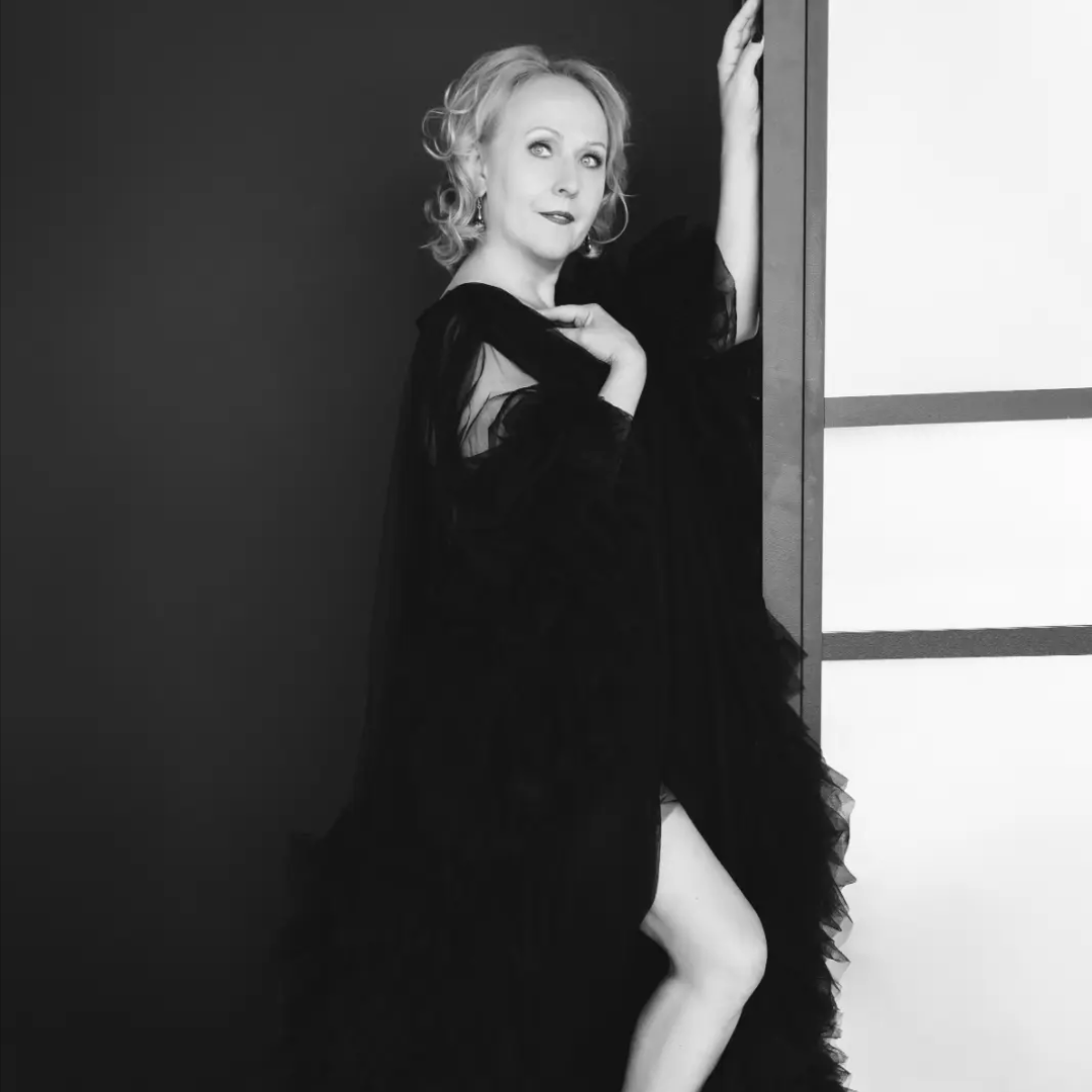 Black and white portrait of a woman with light hair wearing a flowing black dress, posing with one hand on a wall and the other near her chest, looking upward with a contemplative expression.