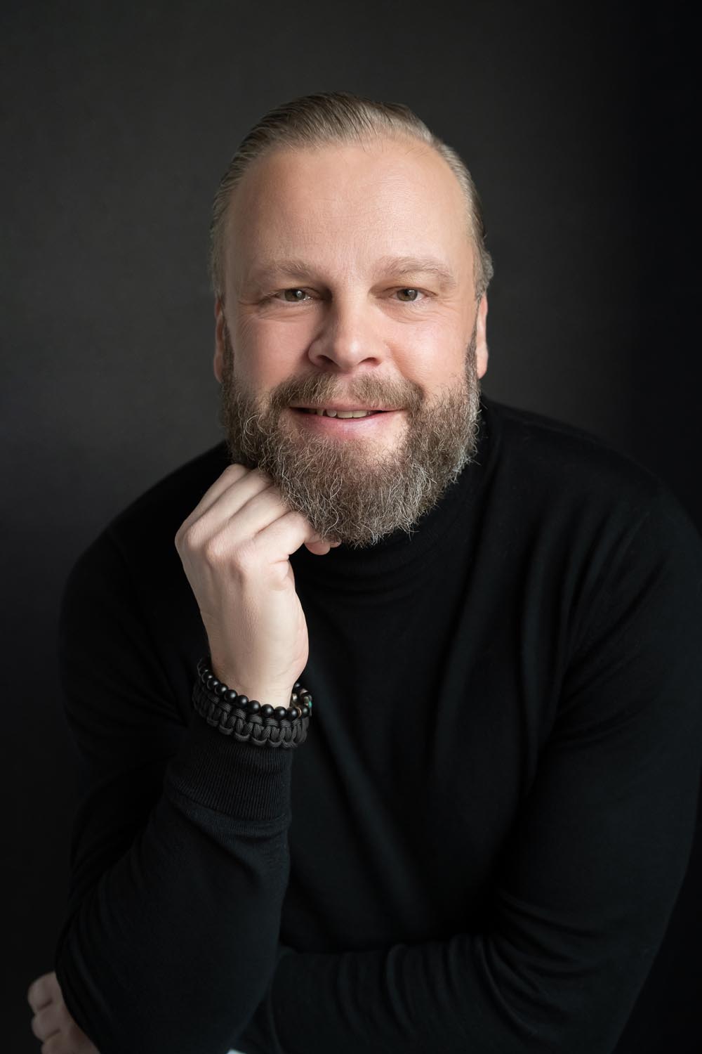 Portrait of a smiling man with a light beard and mustache wearing a black turtleneck, resting his chin on his hand against a dark background.