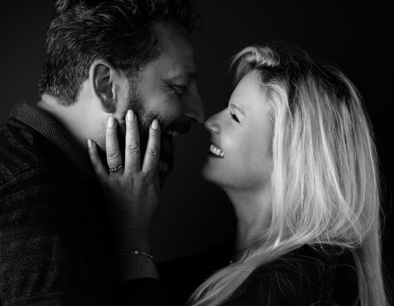 Black and white close-up of a joyful couple facing each other, a woman with long hair smiling and gently holding the man's bearded face.