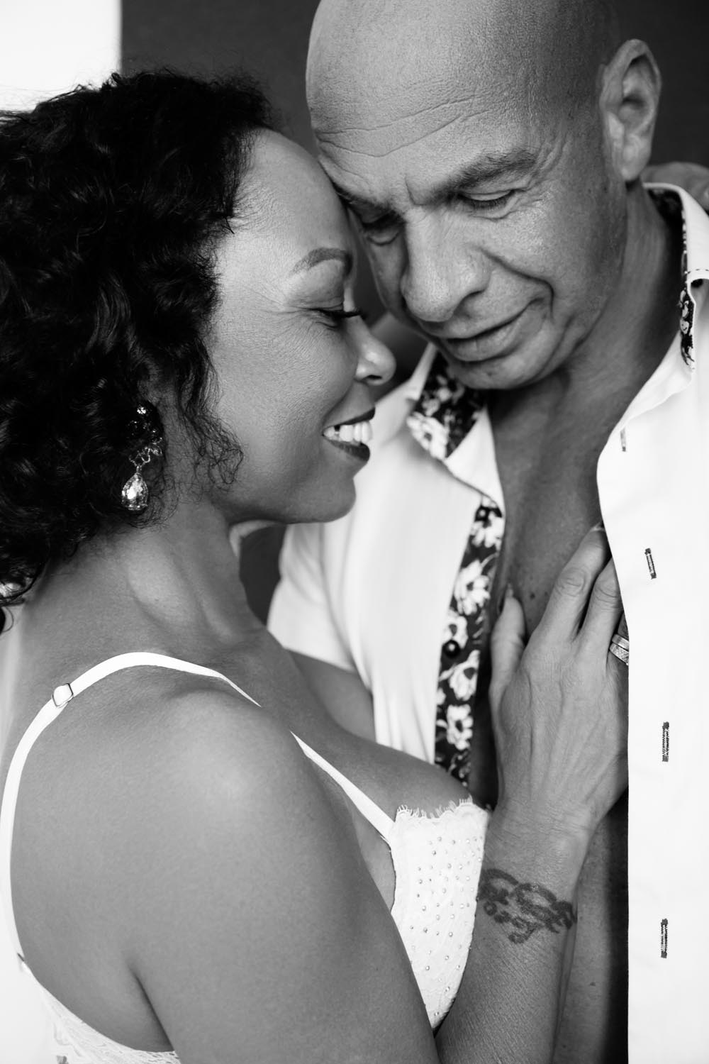 Black and white close-up of a smiling woman and a man with their foreheads together in an intimate pose, the woman wearing earrings and a white top, the man in a partially open shirt, with the woman's hand resting on his chest.
