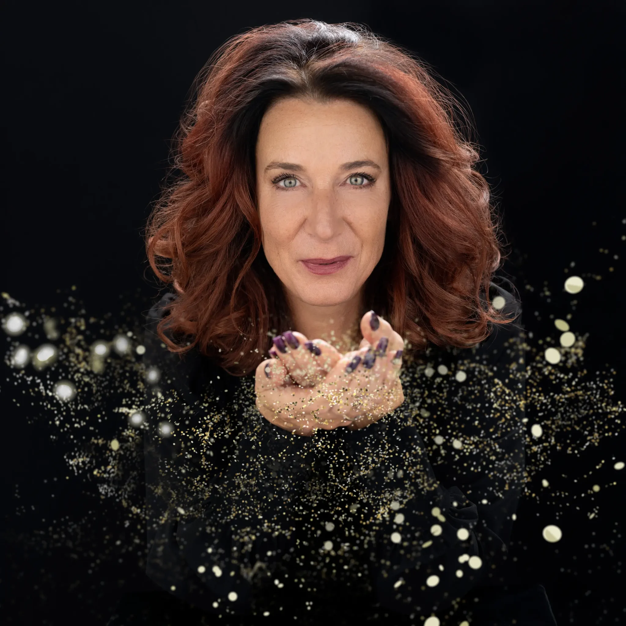 Woman with red curly hair and green eyes blowing gold glitter from her cupped hands against a black background.