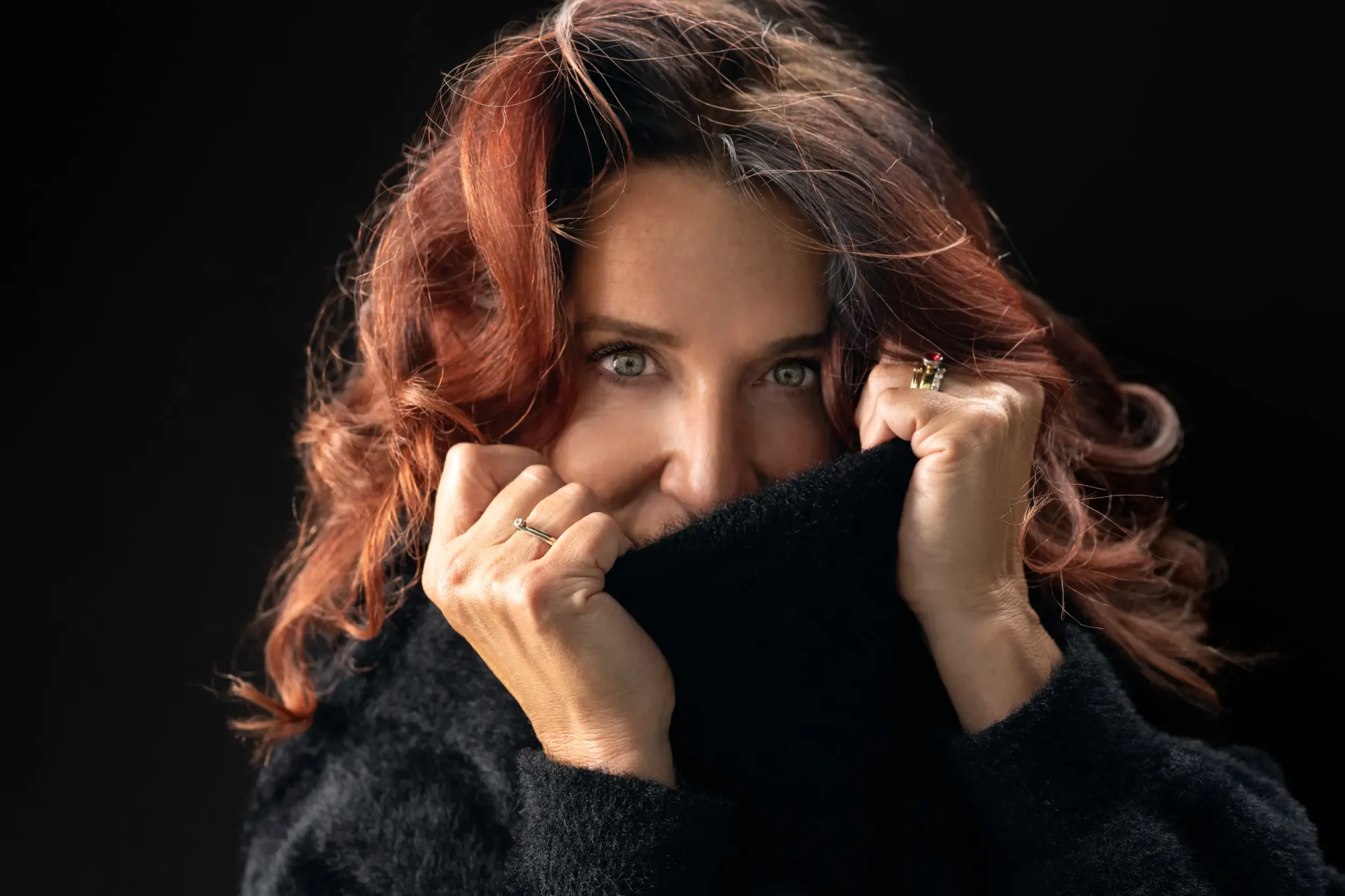 Woman with red curly hair and green eyes wearing a black sweater, pulling the collar up to cover her mouth, against a dark background.