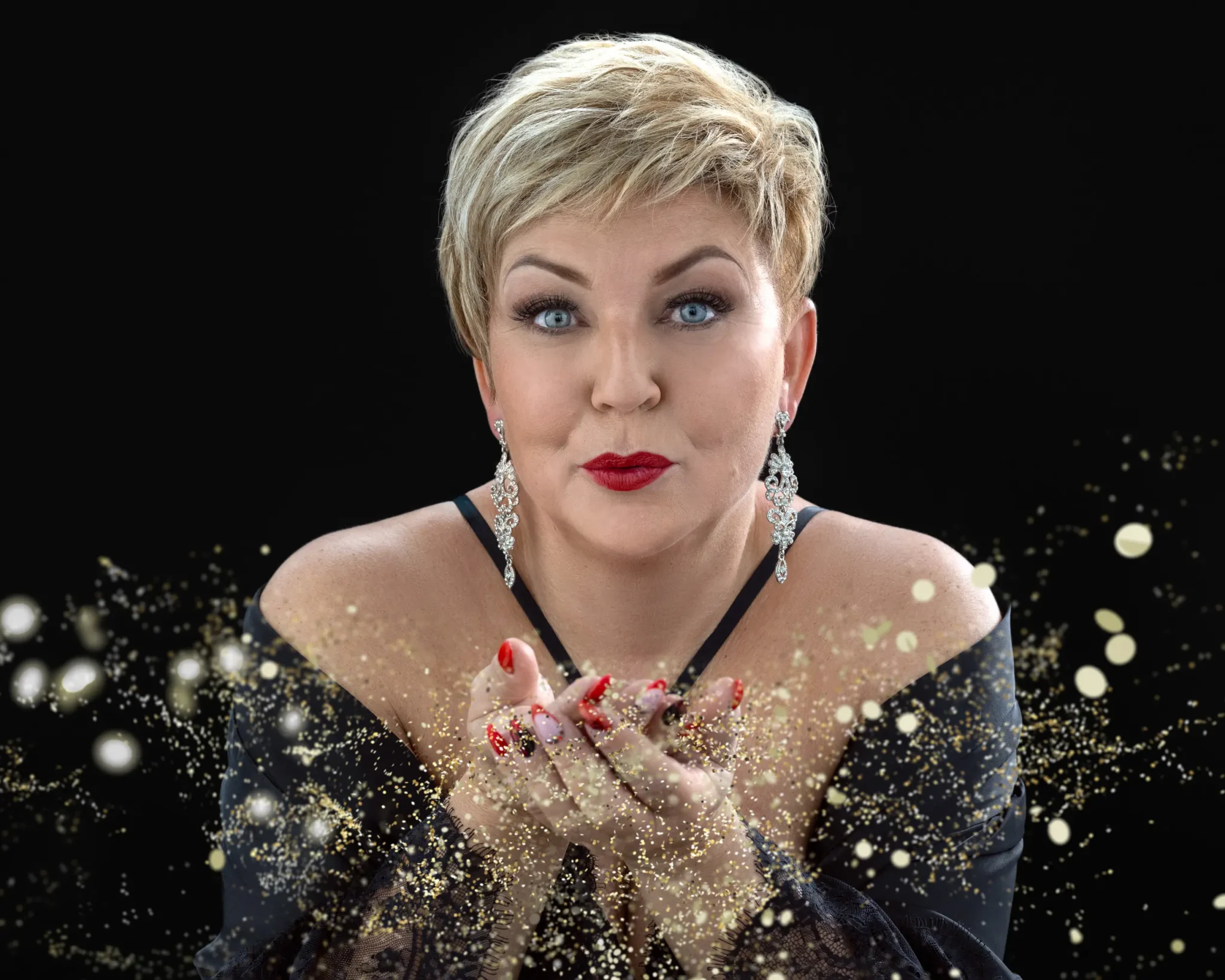 Woman with short blonde hair and blue eyes wearing a black off-shoulder outfit, large silver earrings, and red lipstick, blowing shimmering gold glitter from her cupped hands against a black background.