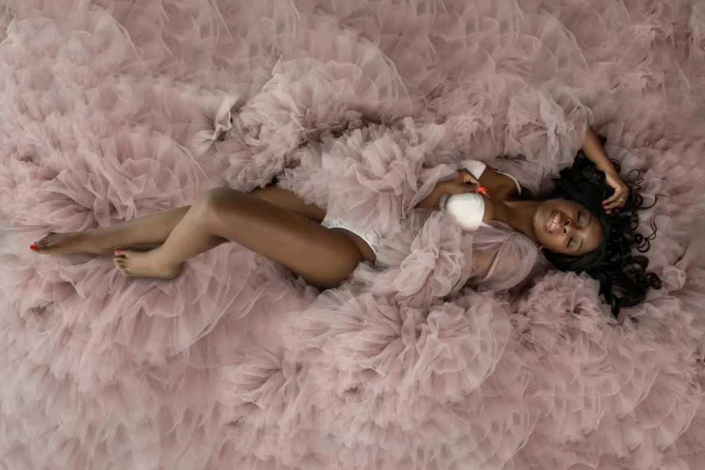 Smiling woman with dark skin and long curly hair lying on a fluffy, voluminous pink tulle fabric, wearing white lingerie and a sheer pink tulle robe.