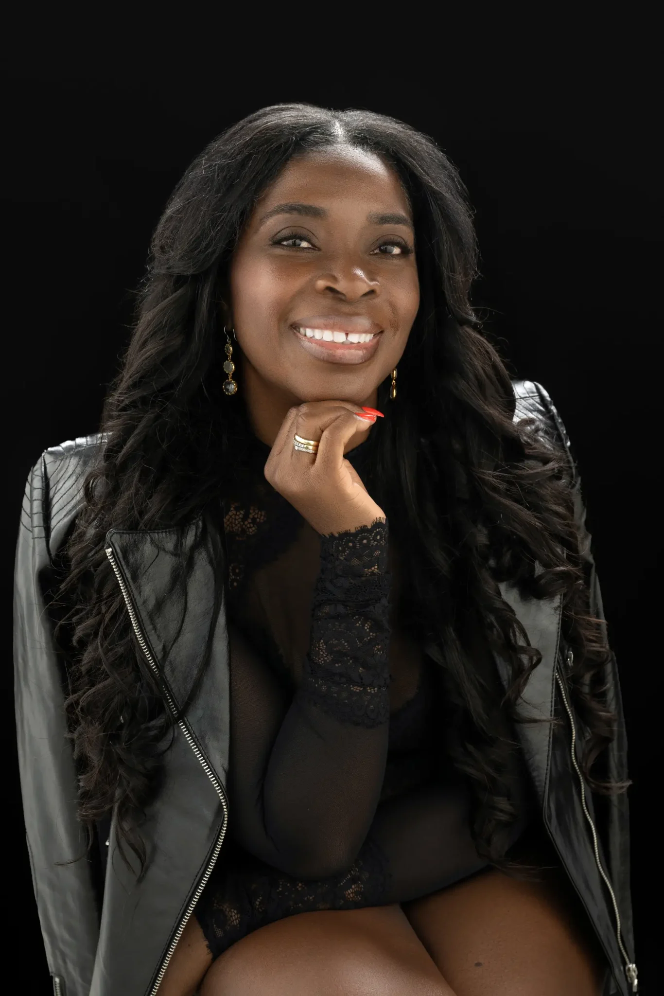Smiling woman with long curly hair wearing a black lace top and a black leather jacket, resting her chin on her hand against a black background.