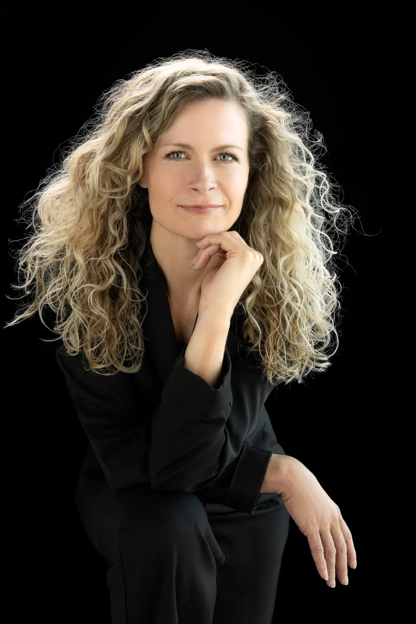 Woman with long curly blonde hair wearing a black outfit, resting her chin on her right hand and looking forward against a black background.