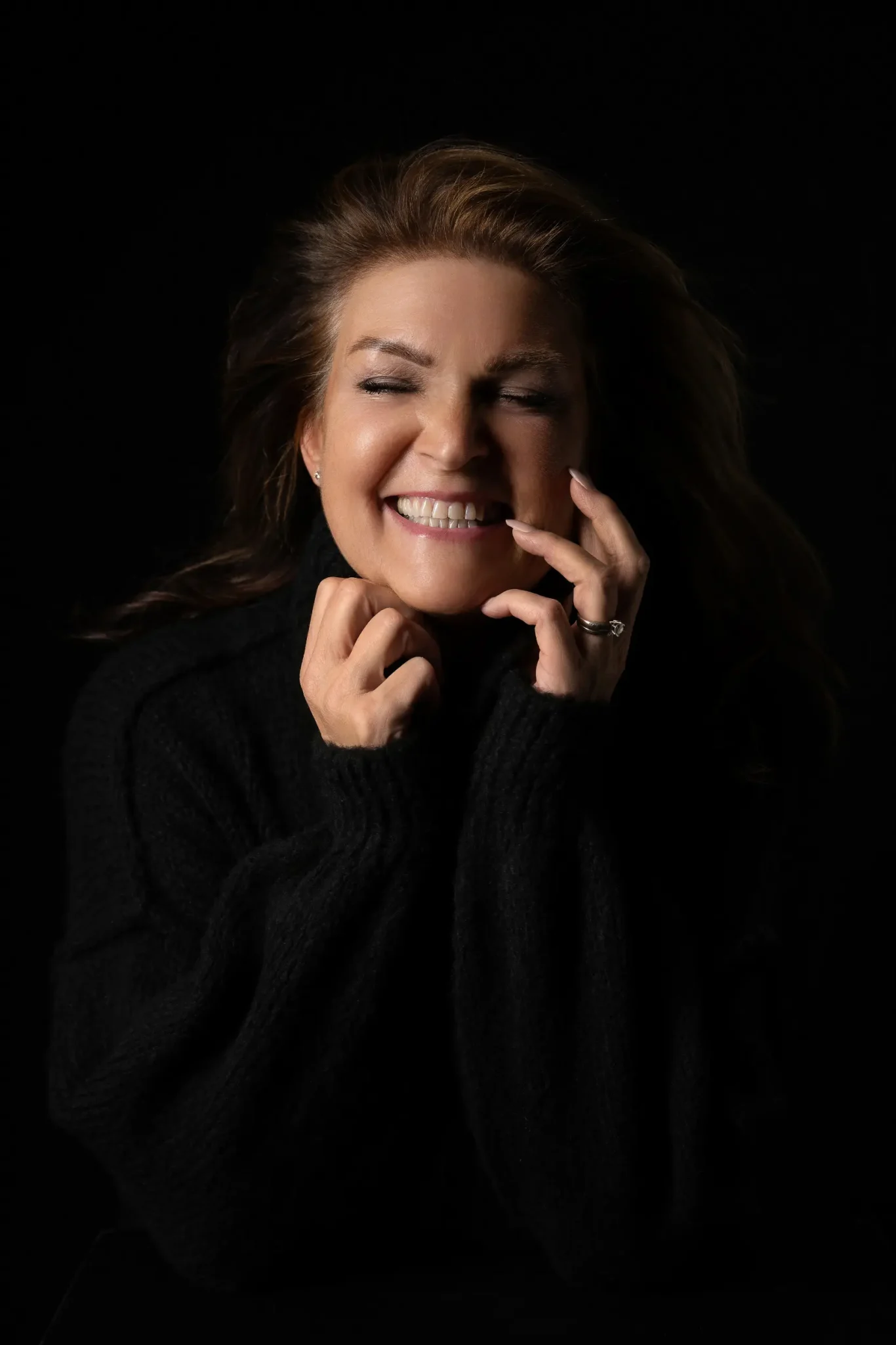 Woman with light brown hair wearing a black sweater smiling with eyes closed and hands touching her face against a black background.