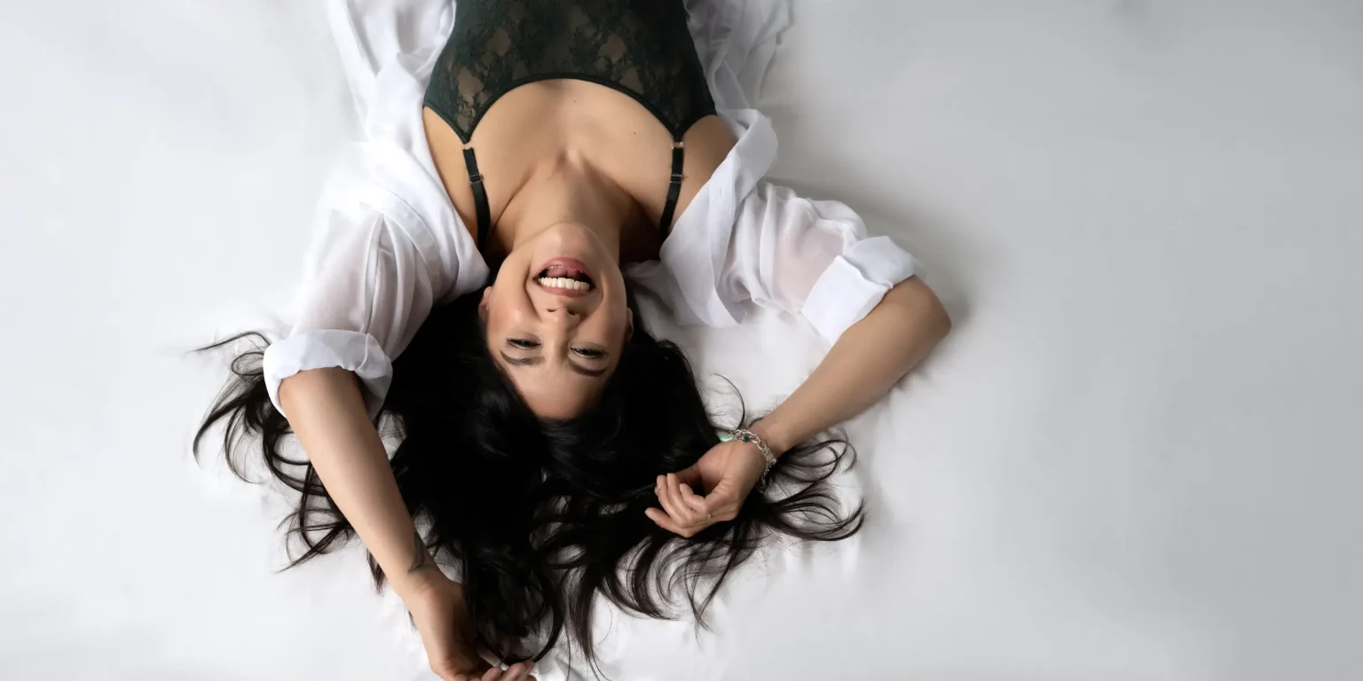 Overhead view of a smiling woman lying on her back on a white surface, wearing a black lace top and a white open shirt, her dark hair spread around her head, and one hand touching her hair.