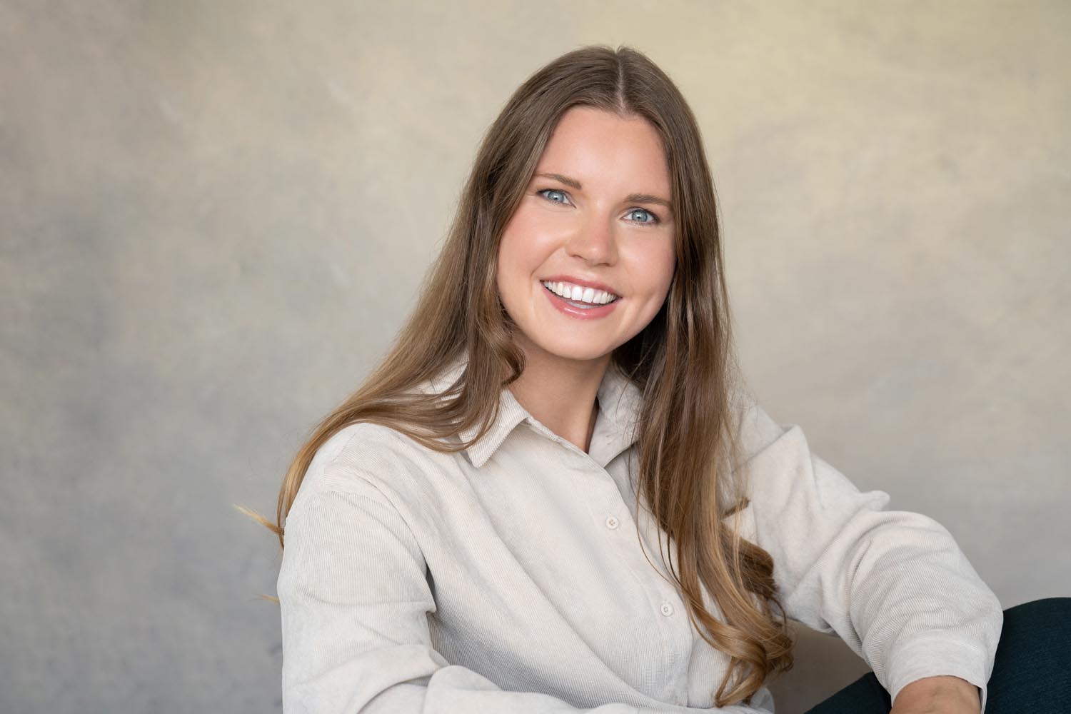Smiling young woman with long light brown hair wearing a beige button-up shirt, sitting against a plain light grey background.