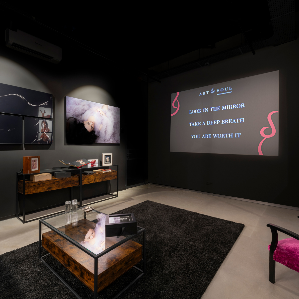 Modern gallery room with dim lighting featuring photographic art on dark gray walls, a wooden and glass coffee table on a black rug, and a wooden console with photo books and framed pictures. On one wall is a projection displaying the text: 'ART & SOUL by Katrina Tromb, LOOK IN THE MIRROR, TAKE A DEEP BREATH, YOU ARE WORTH IT,' accented with red decorative swirls.