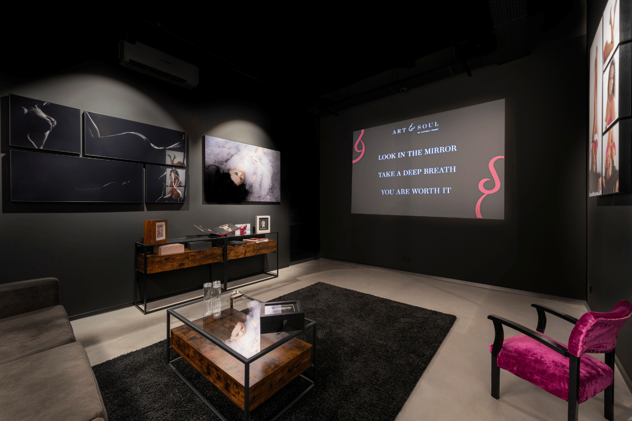 Dark room with modern decor featuring a grey sofa, pink velvet chair, black rug, two wooden tables, and art photographs on the walls, with a projection displaying motivational text: 'LOOK IN THE MIRROR, TAKE A DEEP BREATH, YOU ARE WORTH IT' under the heading 'ART & SOUL'.