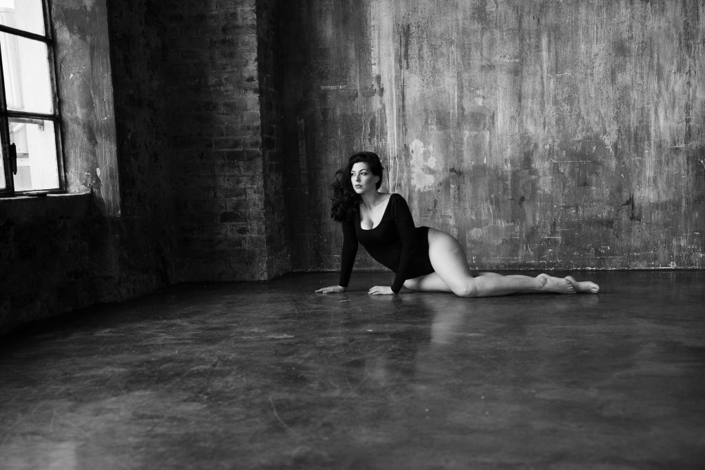Black and white photo of a woman in a long-sleeve black leotard lying on the floor of a spacious, industrial-style room with textured concrete walls and large windows.