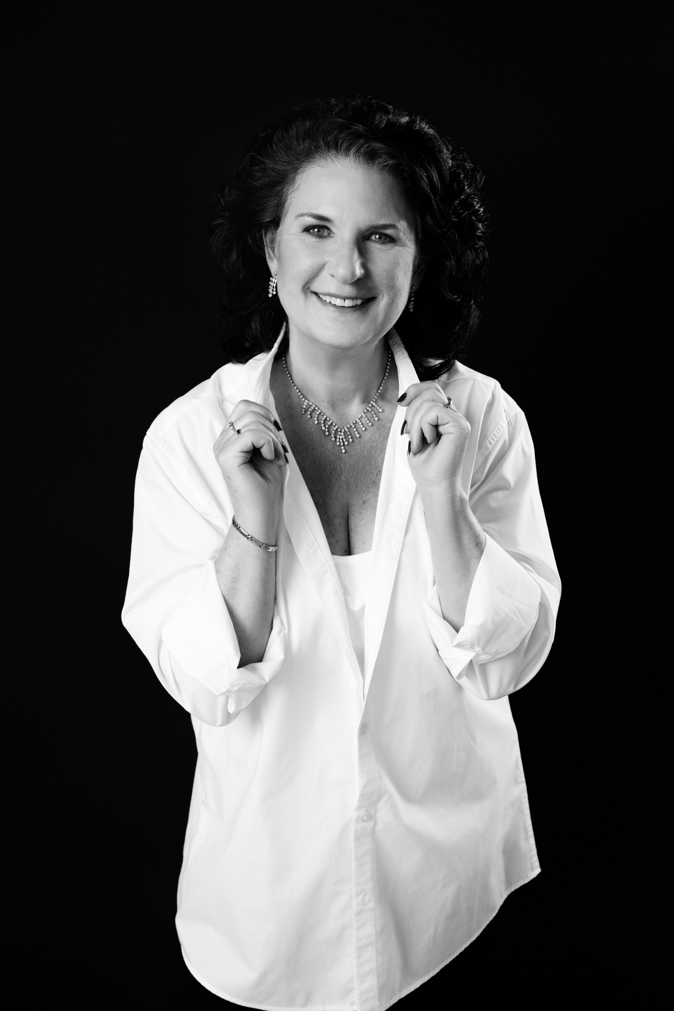 Smiling middle-aged woman with dark curly hair wearing a white oversized button-down shirt, diamond necklace, and matching earrings against a black background.