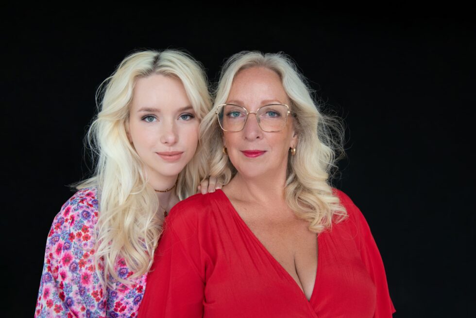 Two blonde women with light skin posing against a black background; the younger woman wears a floral top and the older woman in front wears glasses and a red blouse.