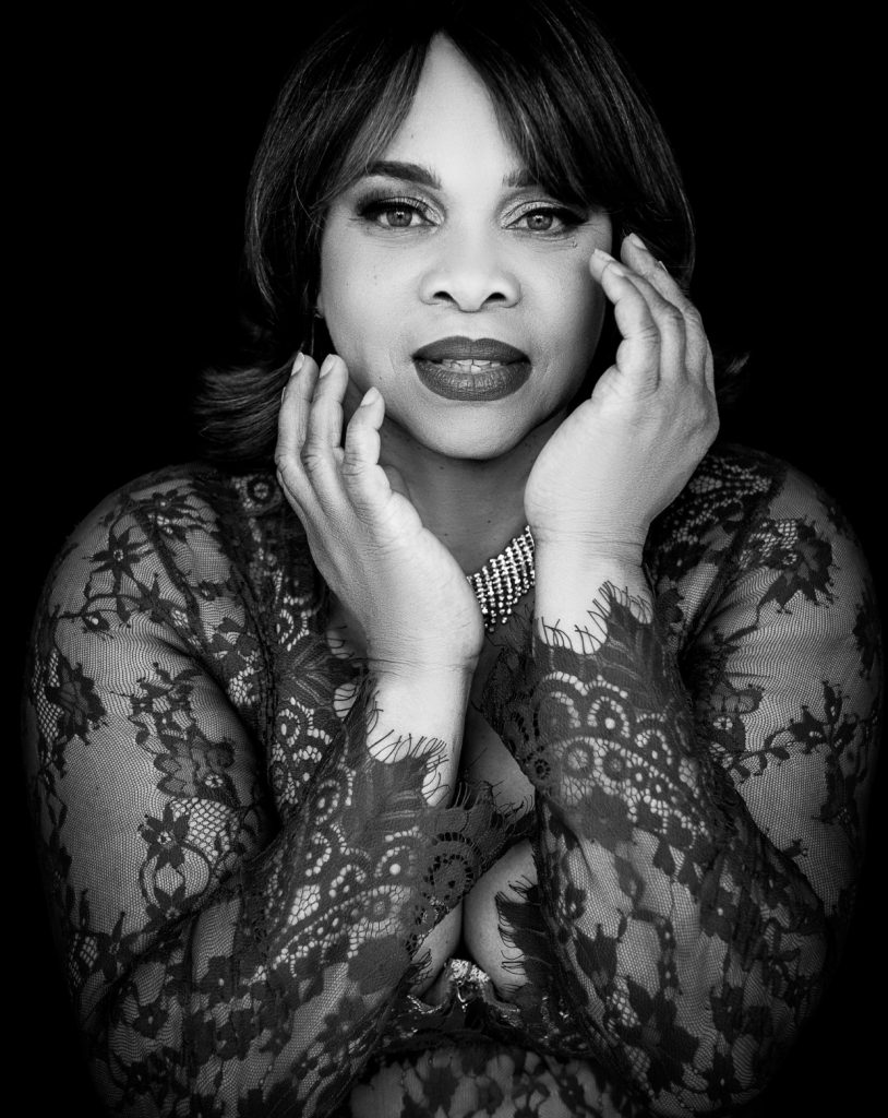 Black and white portrait of a woman with shoulder-length hair wearing a lace garment and a statement necklace, posing with hands gently framing her face against a black background.