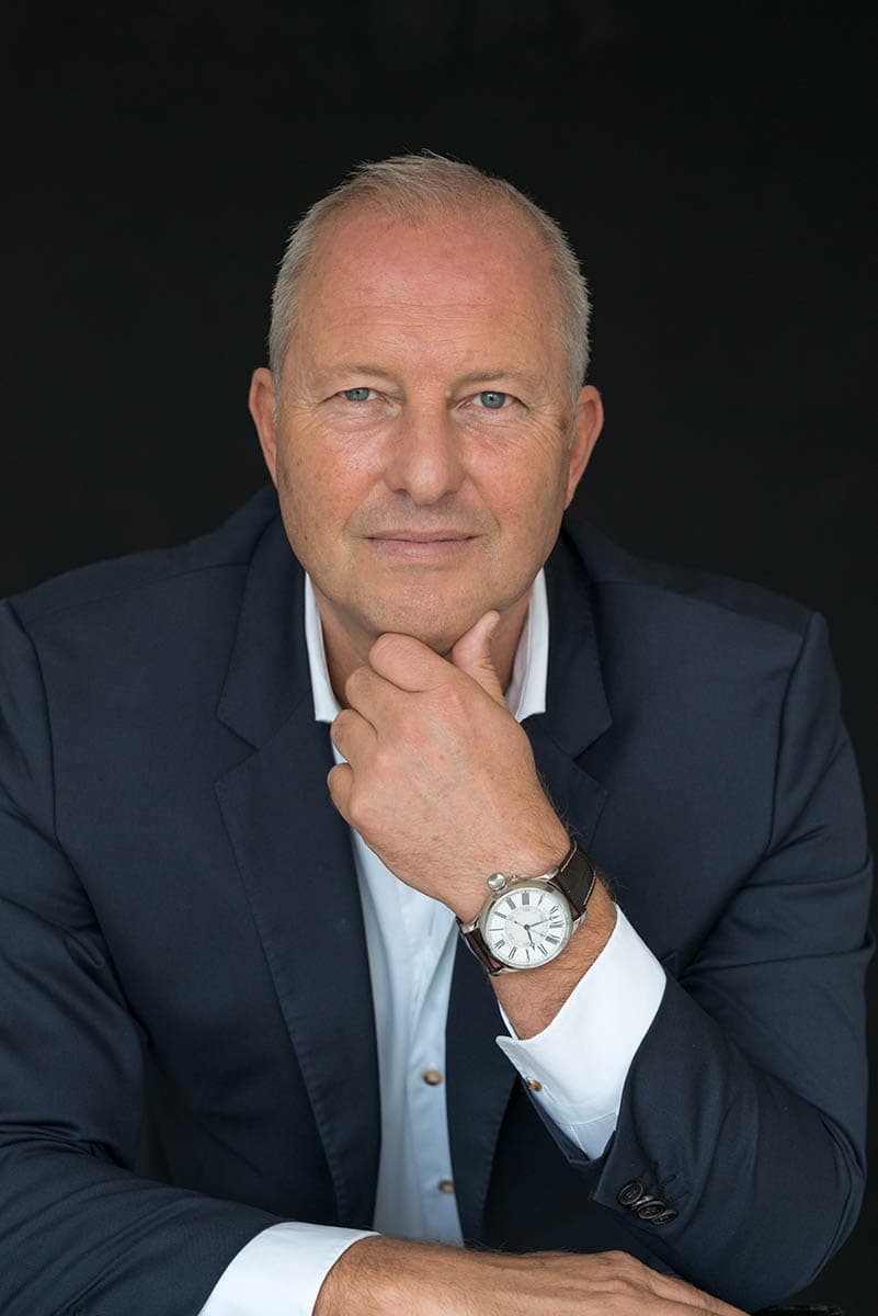 Middle-aged man with short gray hair wearing a dark blazer and white shirt, resting his chin on his hand showing a wristwatch, looking directly at the camera against a black background.