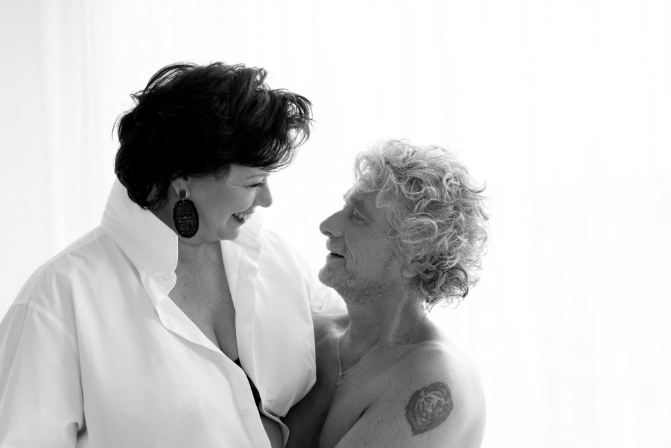 Black and white image of a smiling woman in a white shirt looking lovingly at a shirtless man with curly hair and a tattoo on his upper arm, both sharing an intimate moment.