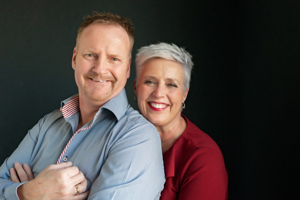 Smiling middle-aged couple posing against a dark background; the man wears a blue shirt with folded arms, and the woman in a red top stands behind him with her arms around his shoulders.