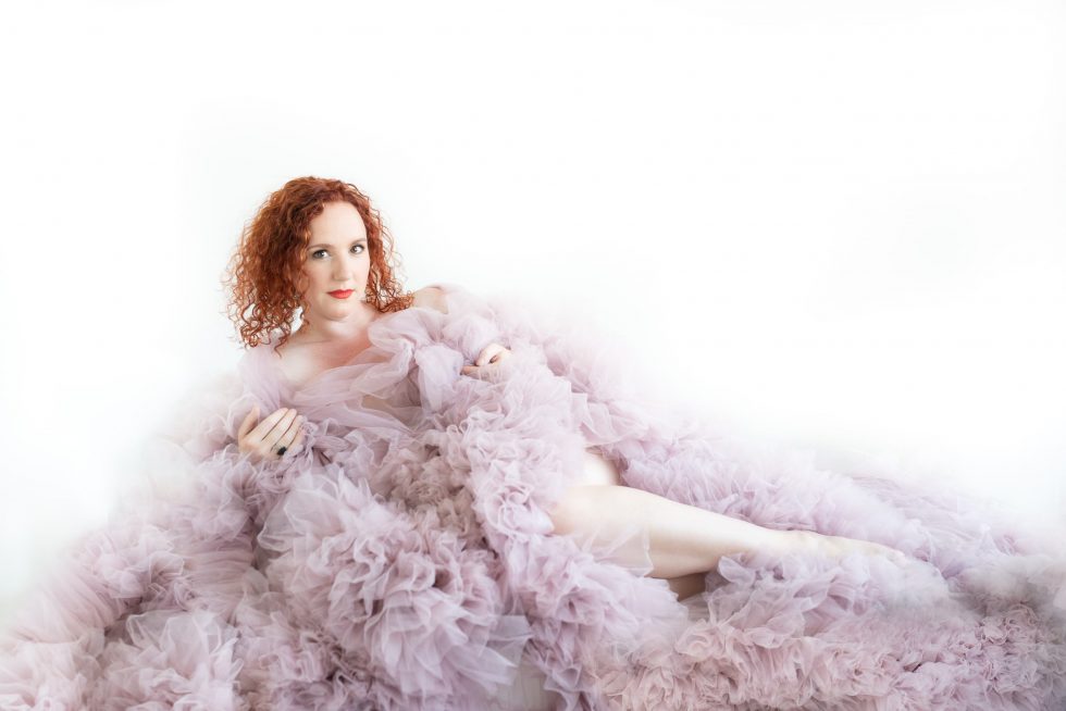 Red-haired woman with curly hair reclining on a white surface, wrapped in voluminous, soft lavender tulle fabric that envelops her body and extends around her.