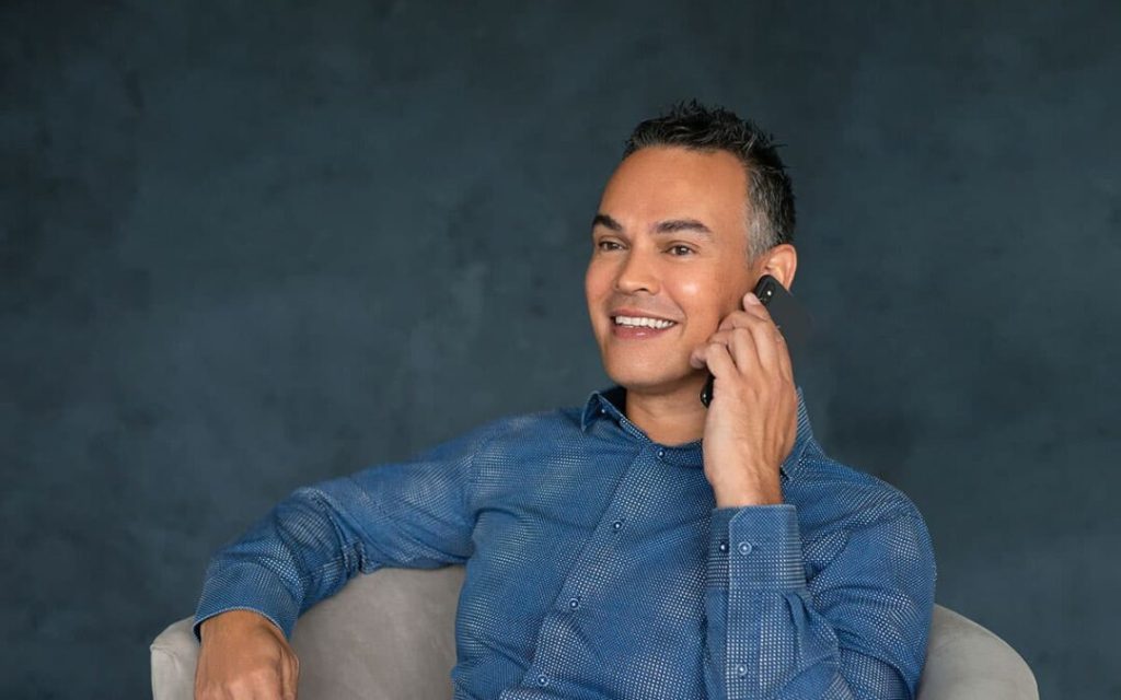 Smiling man sitting on a light gray chair while talking on a mobile phone against a dark gray background.