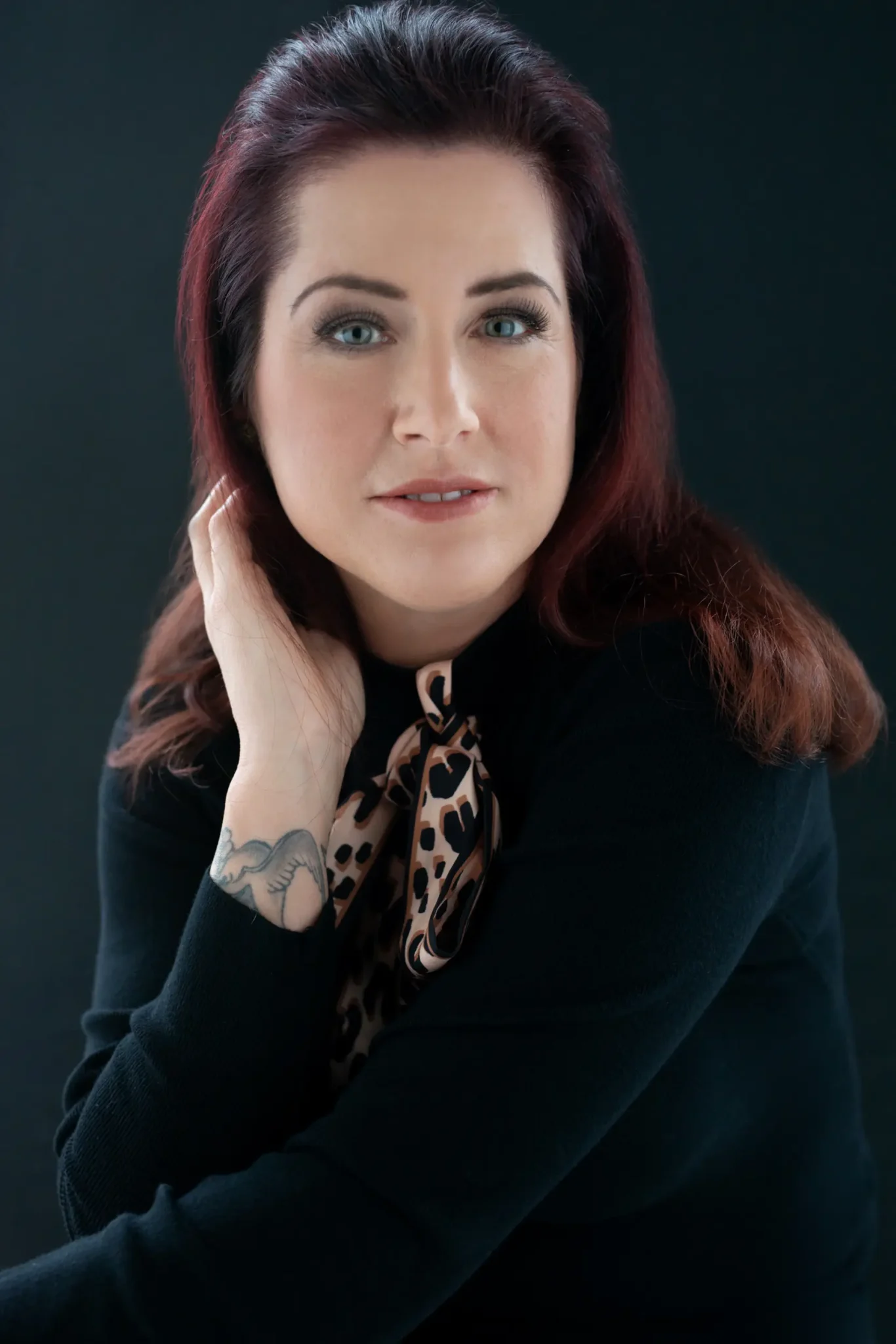 Portrait of a woman with reddish-brown hair wearing a black sweater and leopard print scarf, resting her hand on her face, showing a tattoo of a bird on her wrist, against a dark background.
