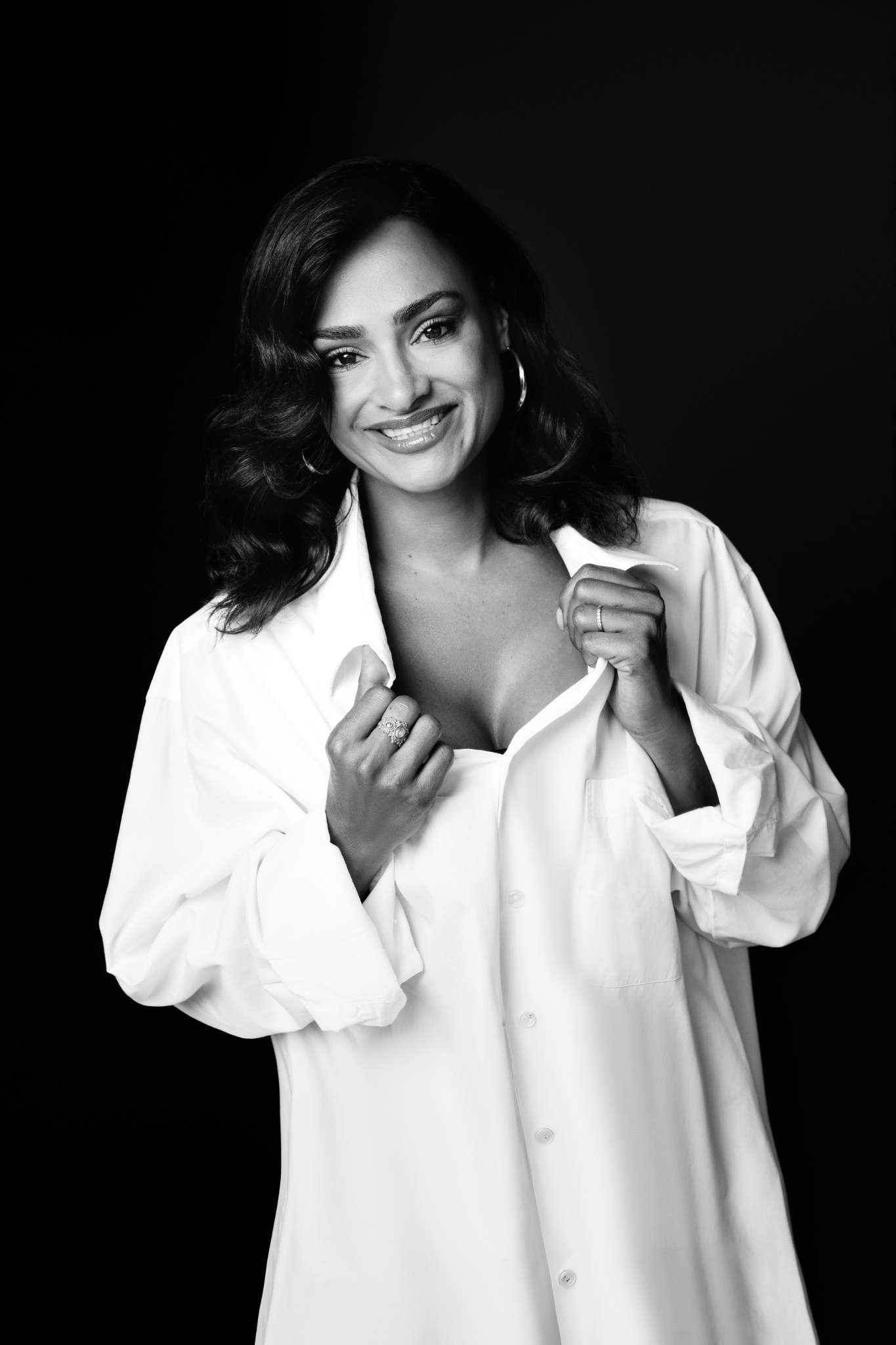 Black and white portrait of a smiling woman with wavy hair wearing a loose white button-up shirt, holding the collar with both hands against a dark background.