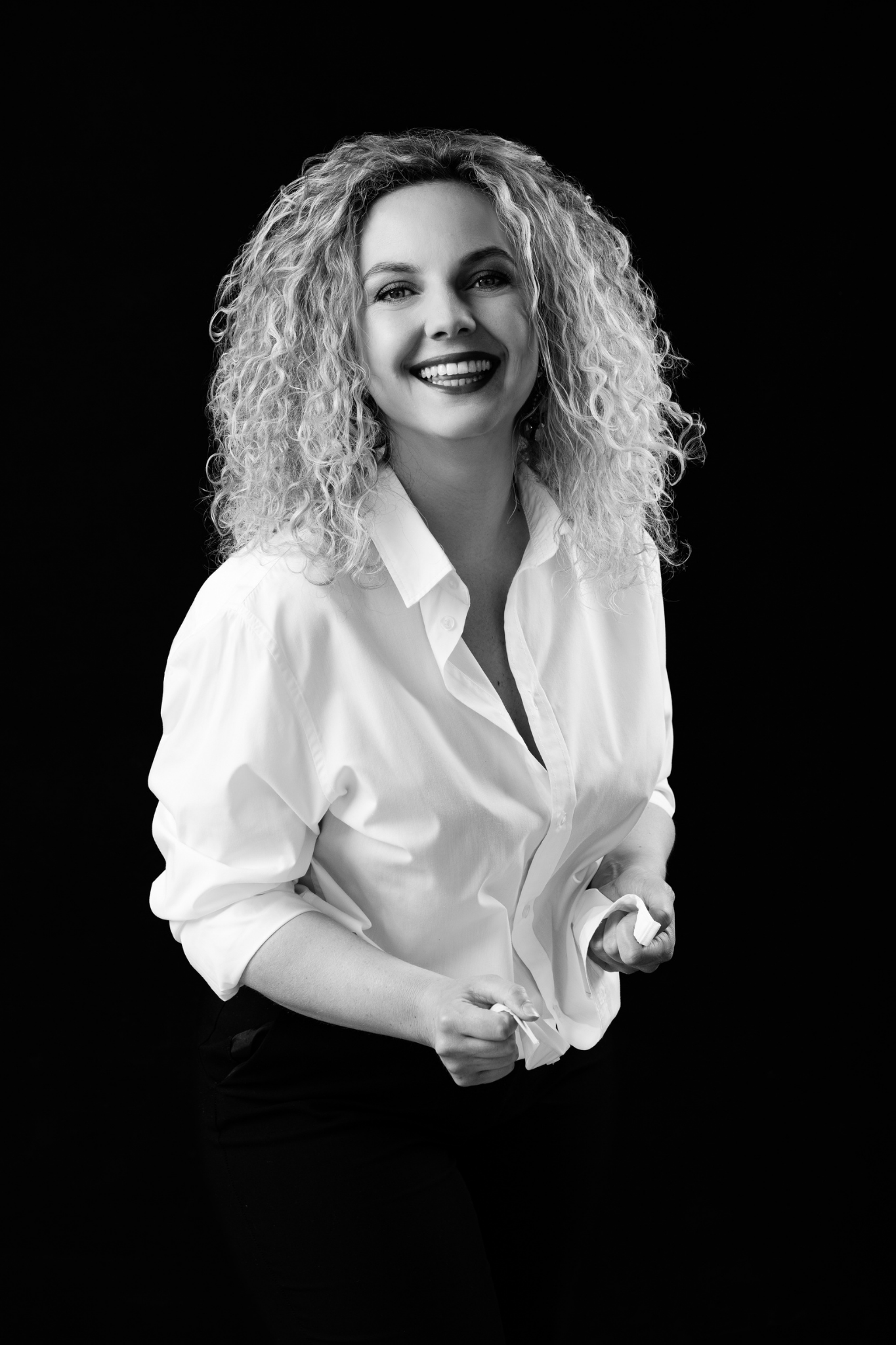 Black and white portrait of a smiling woman with curly hair wearing a white button-up shirt and black pants, standing against a black background.
