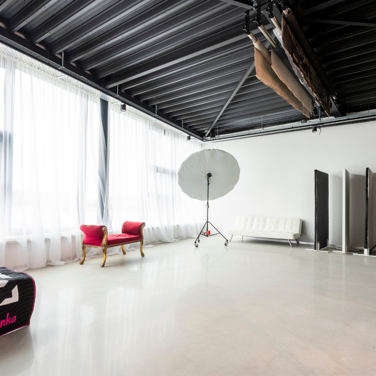 Bright, spacious photography studio with large sheer curtains along one wall, a white umbrella light reflector on a stand, a red and gold vintage bench, a white modern sofa, and black and white photography reflectors standing against a white wall under a black industrial ceiling with hanging fabric backdrops.