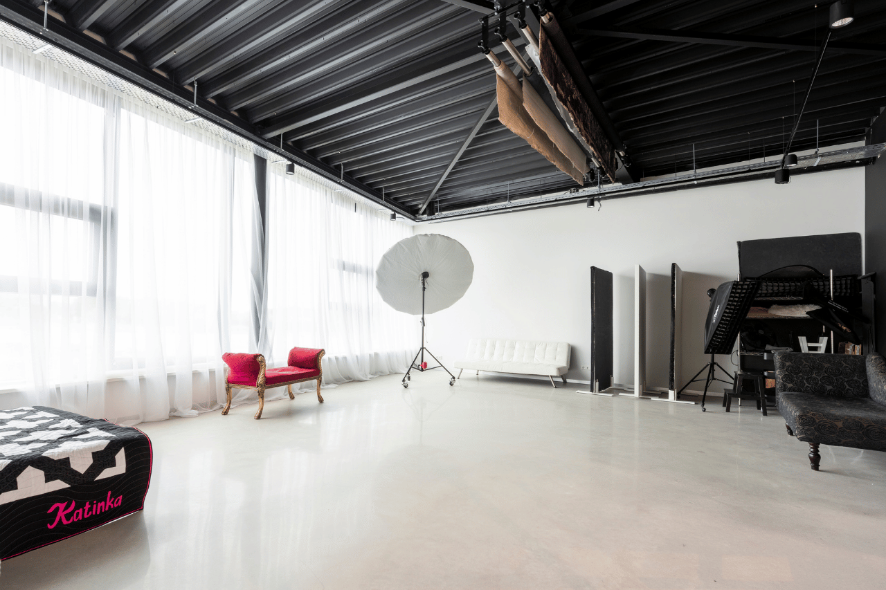 Bright photography studio with large windows and sheer white curtains, featuring a red vintage bench, a white modern sofa, a black patterned sofa, photography lighting equipment including a large umbrella light on a stand, and various reflectors and backdrops.