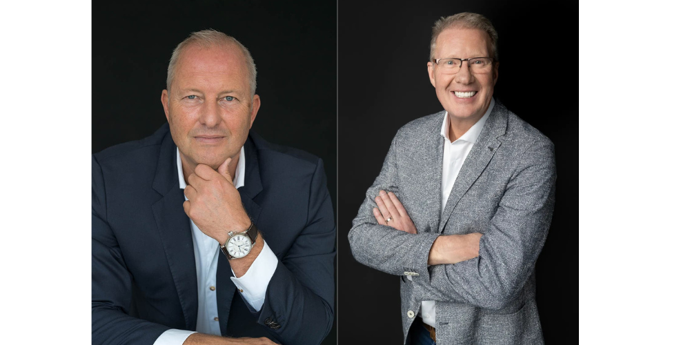 Two professional men posing against a black background; the man on the left wears a dark blazer and watch, resting his chin on his hand with a neutral expression, the man on the right smiles broadly, wearing glasses and a grey blazer with arms crossed.