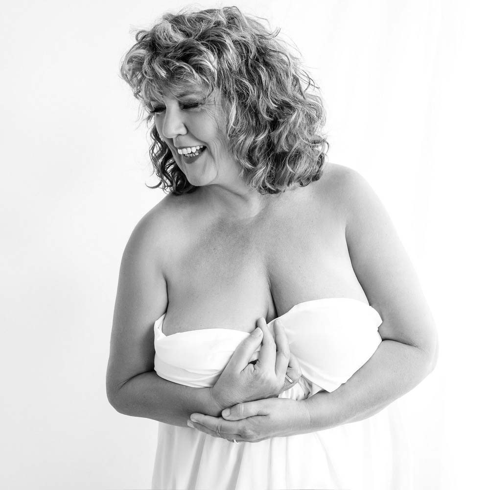 Black and white photo of a smiling woman with curly hair wrapped in a white fabric, holding it at her chest, looking to the side.