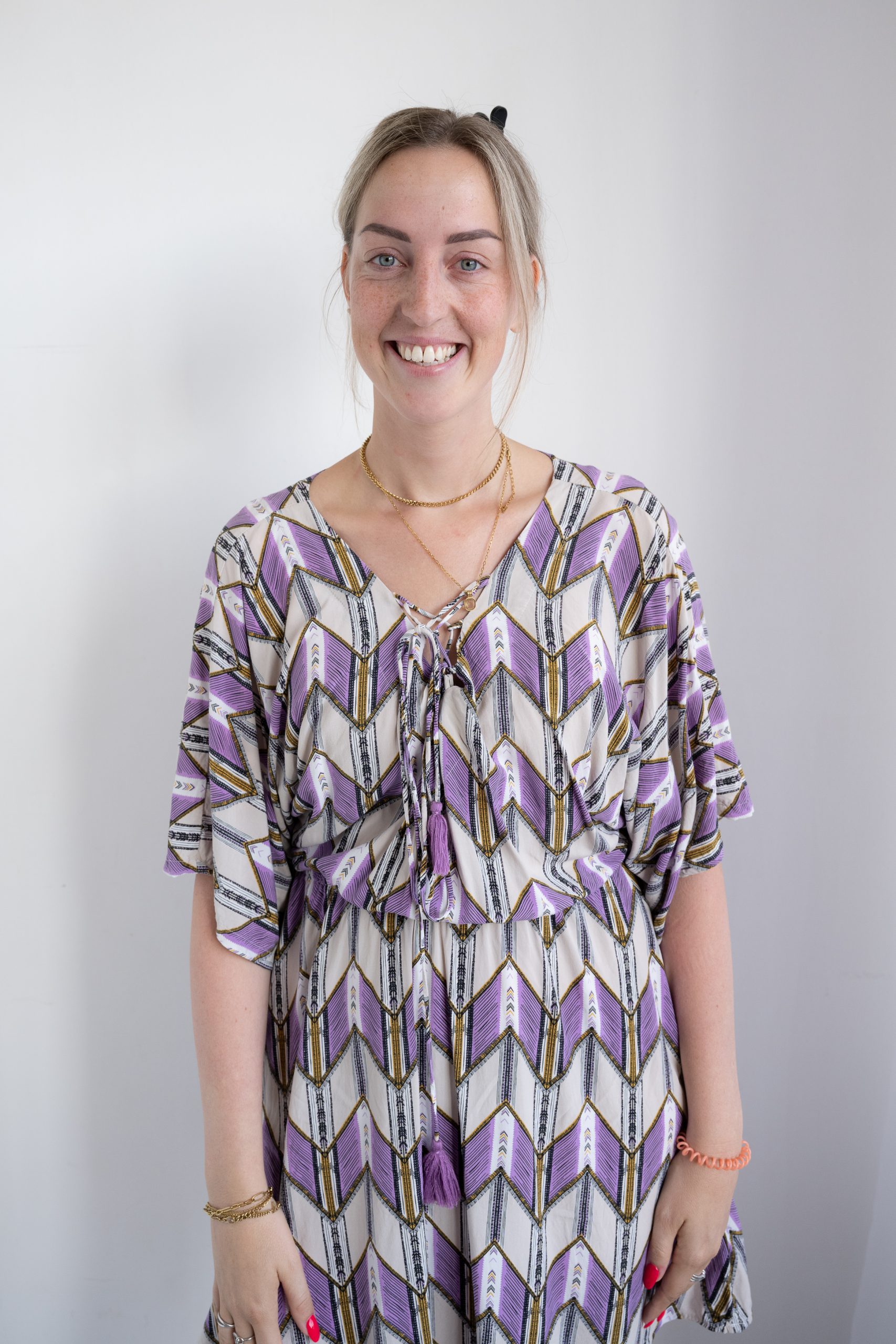 Young woman with light hair tied back, smiling at the camera, wearing a patterned dress with purple, white, black, and gold zigzag designs, and gold necklaces, standing against a plain light gray background.