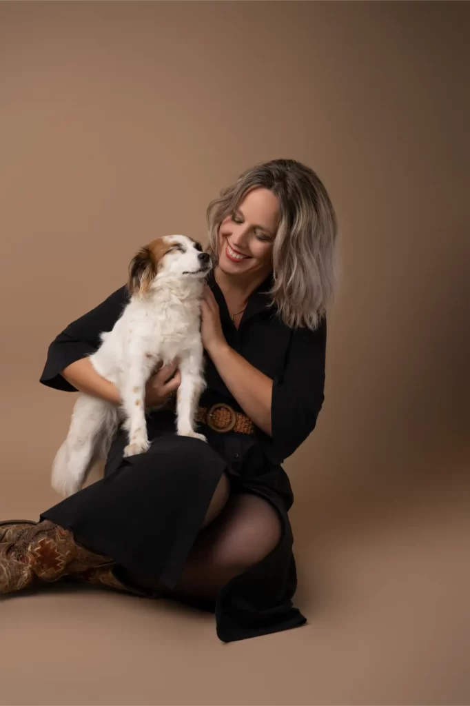 A woman with shoulder-length blonde hair wearing a black dress and brown boots sitting on the floor, smiling and holding a white and brown dog affectionately against a beige background.