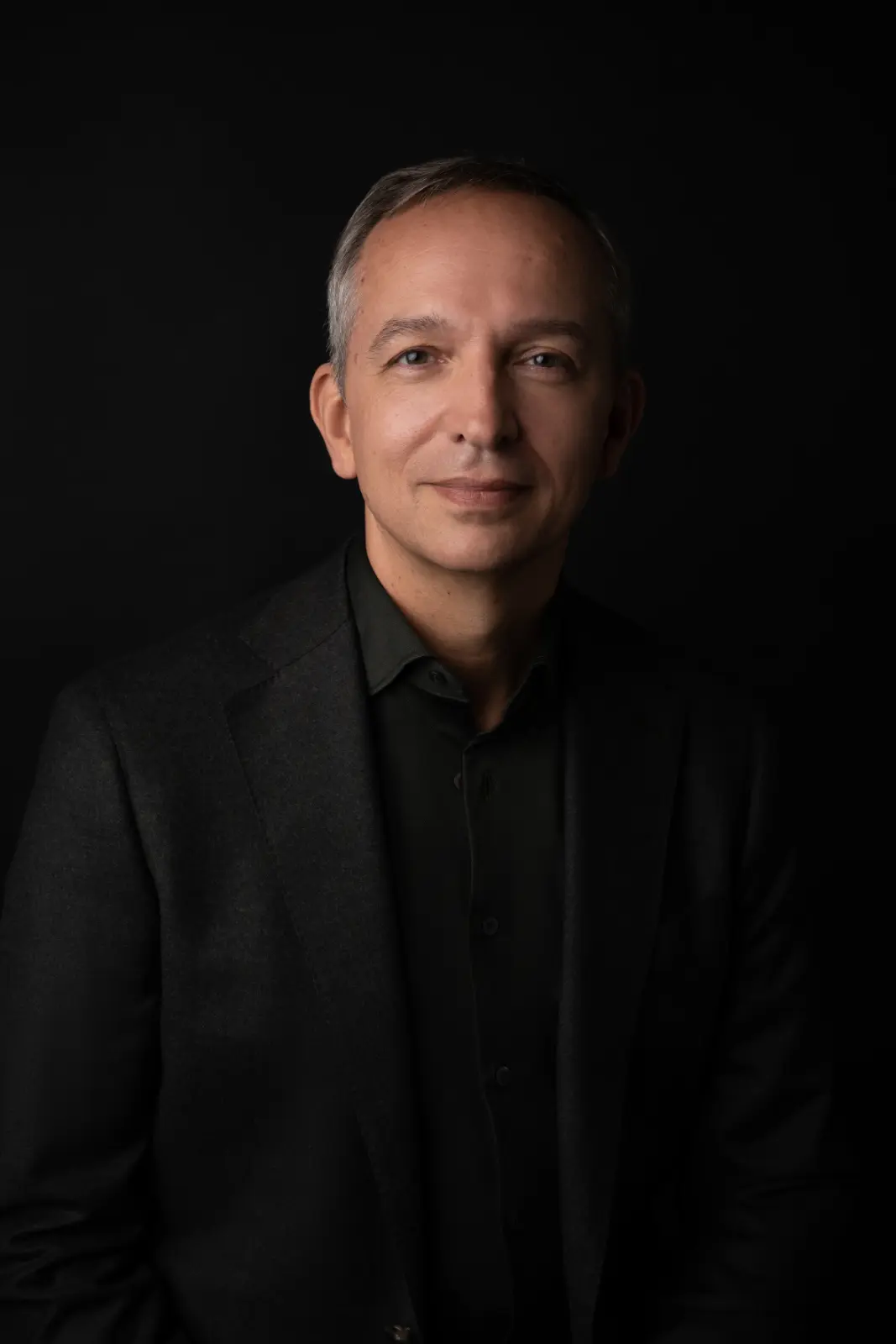 Portrait of a middle-aged man with short gray hair wearing a dark blazer and black shirt against a black background.