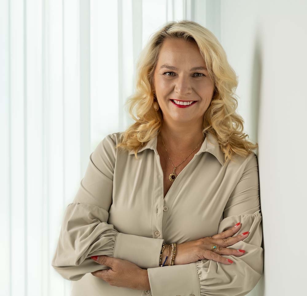 Smiling woman with blonde curly hair leaning against a white wall wearing a beige blouse, red lipstick, and bracelets.