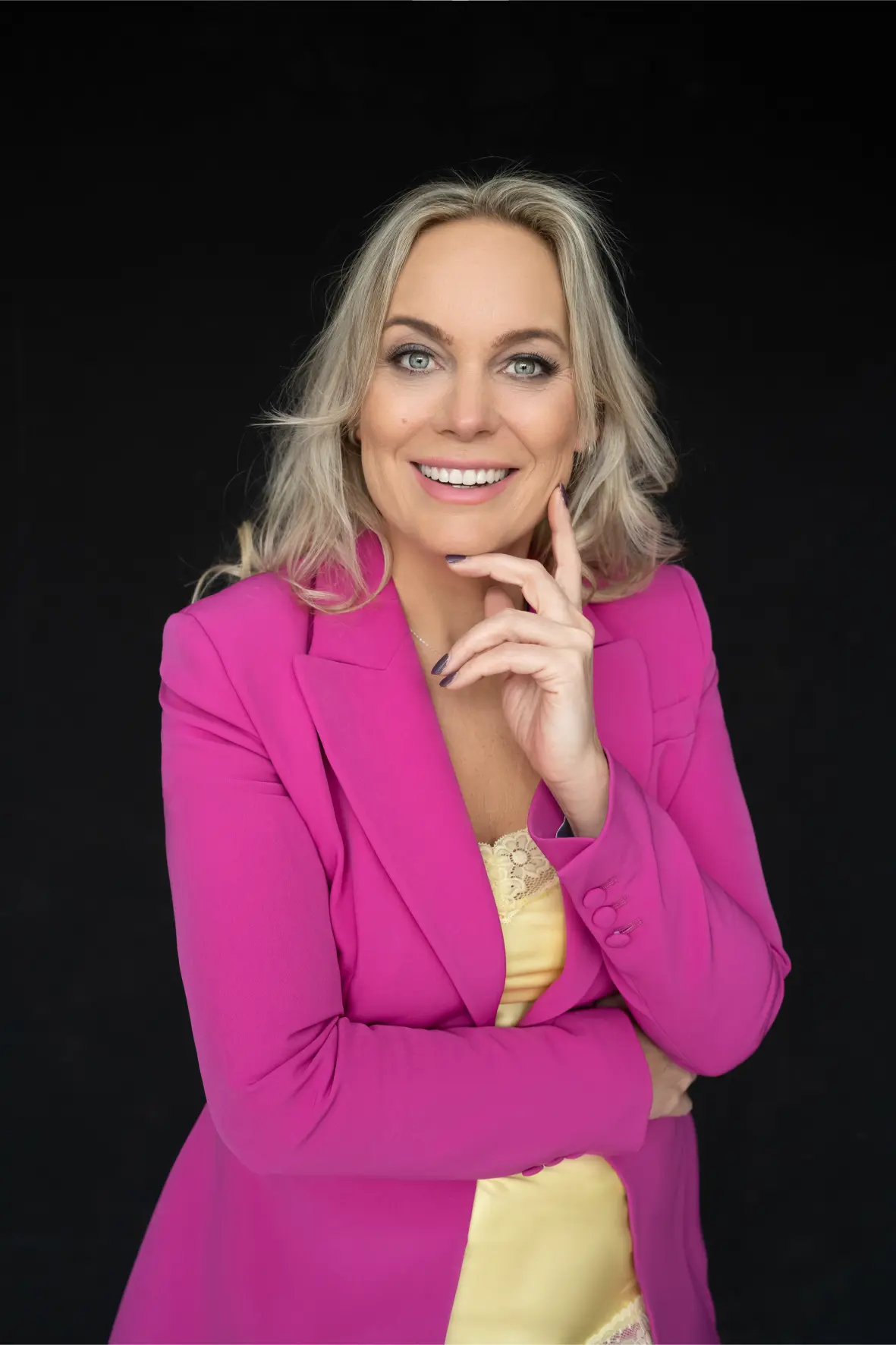 Smiling woman with blonde hair wearing a bright pink blazer over a yellow lace-trimmed top against a black background.