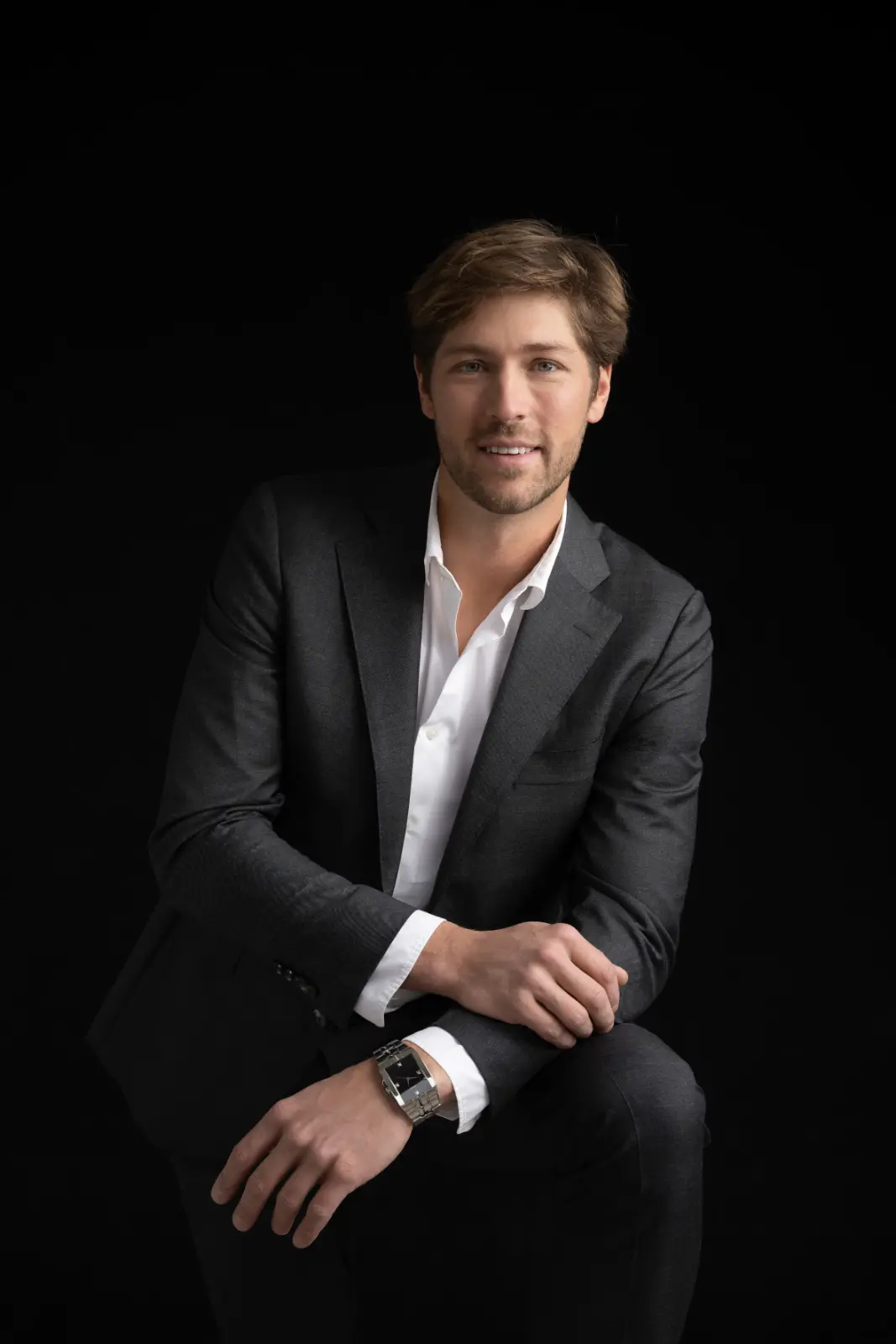A man with light brown hair and a trimmed beard wearing a dark gray suit and white shirt seated against a black background, smiling gently, with one arm resting on his knee and wearing a silver wristwatch.