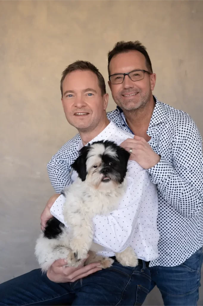 Two men, one wearing glasses, smile and pose together with a small black and white fluffy dog sitting on the lap of the man in front, against a plain beige background.