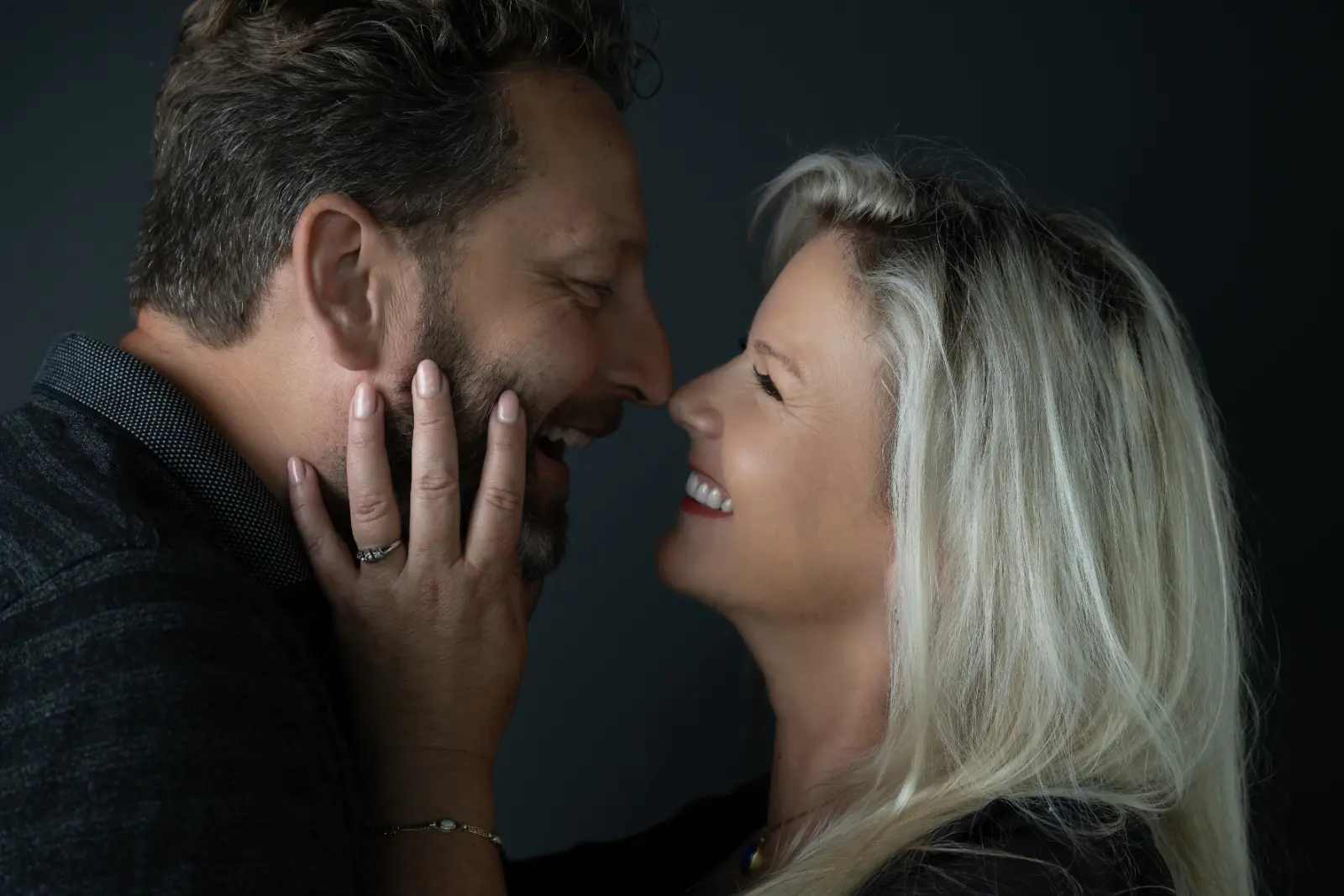 Close-up of a man and a woman smiling and facing each other closely, with the woman's hand gently touching the man's cheek showing a ring on her finger against a dark background.