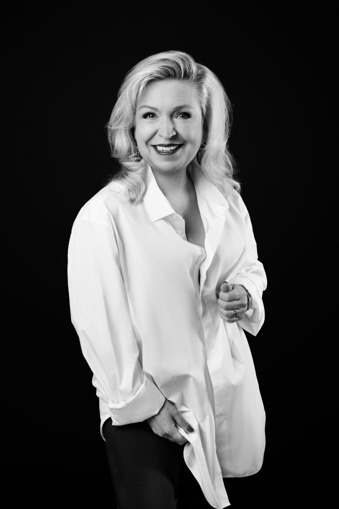 Black and white portrait of a smiling blonde woman wearing a loose white button-up shirt and dark pants, standing against a black background.