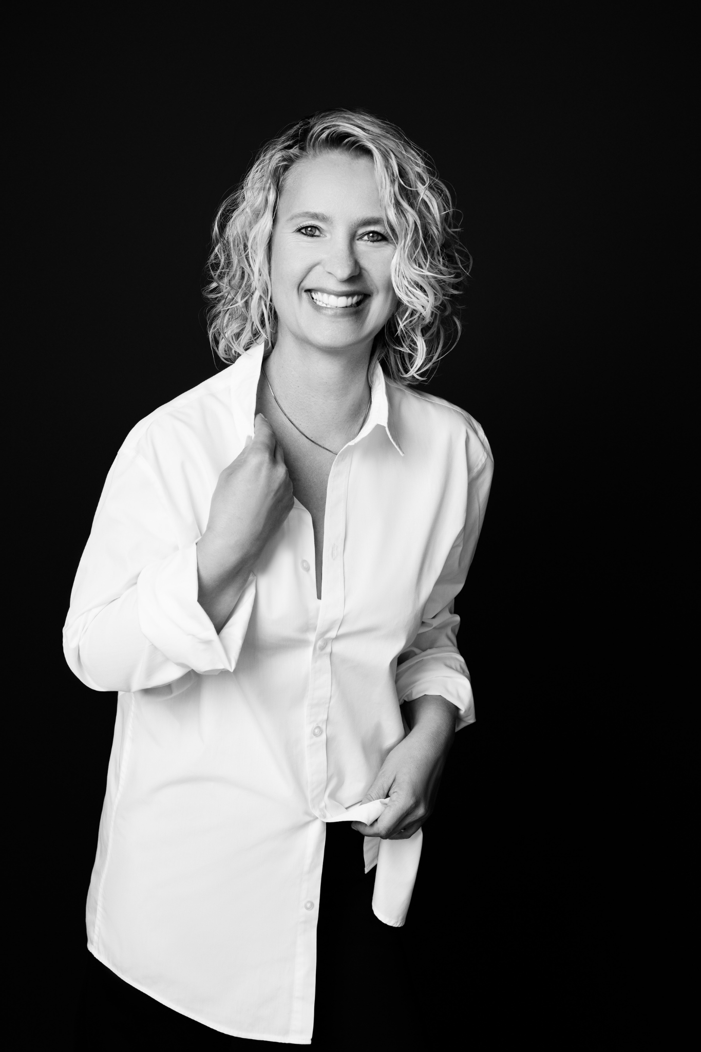 Black and white portrait of a smiling woman with curly hair wearing a white button-up shirt against a black background.
