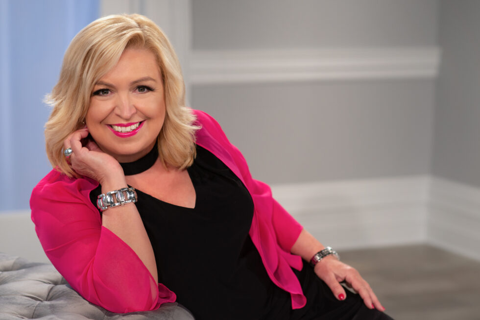 Smiling blonde woman wearing a bright pink shawl over a black top, reclining on a gray tufted sofa with her head resting on her hand, against a softly lit neutral indoor background.