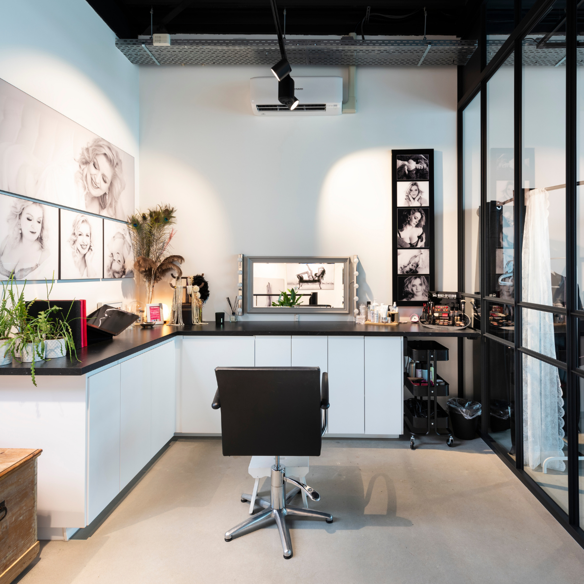 Modern makeup studio with a black swivel chair facing a black countertop with white cabinets, a large vanity mirror, and makeup products, framed black-and-white portraits on walls, glass partition on the right, and decorative plants on the left.