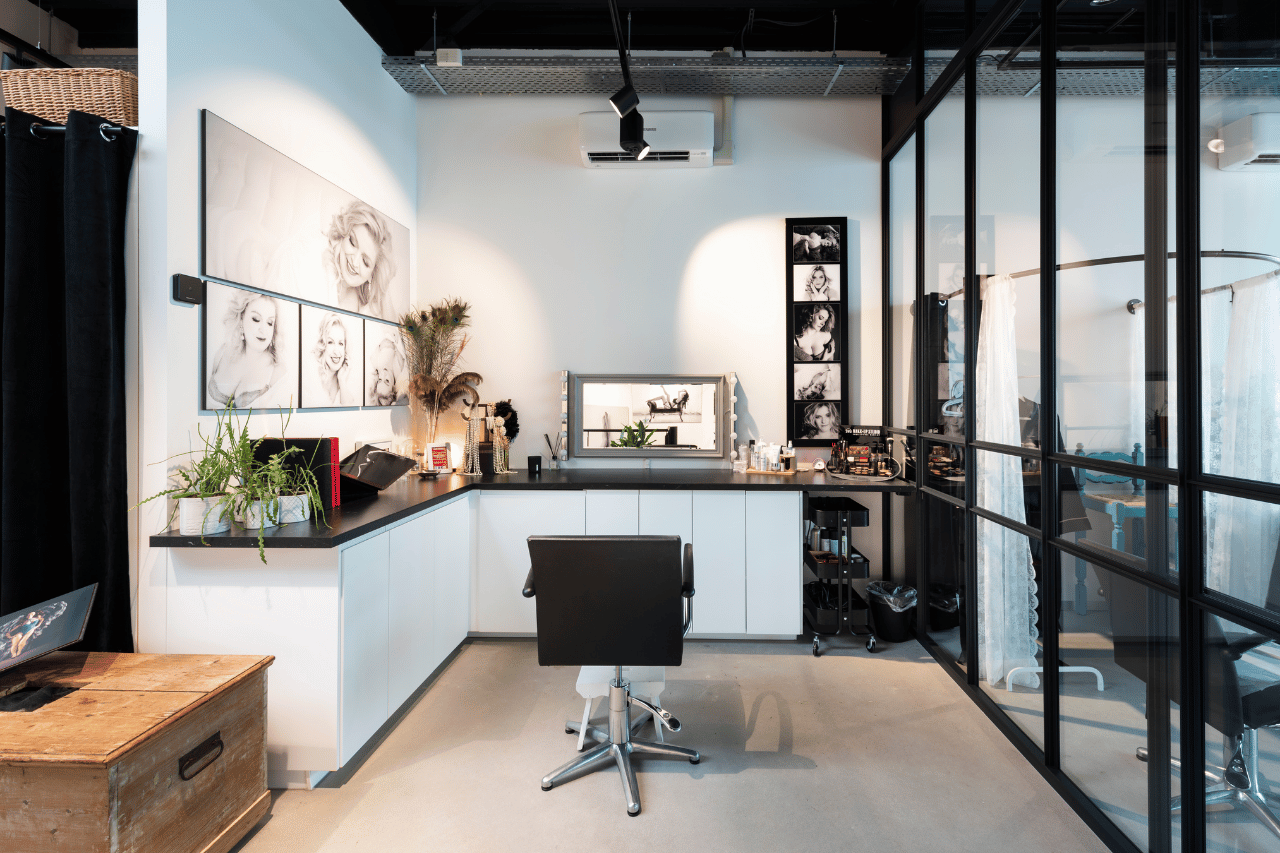 Modern makeup studio with a black swivel chair in front of a vanity mirror surrounded by makeup products and black-and-white portrait photos on the walls, plants on the counter, and glass partition dividing the space.